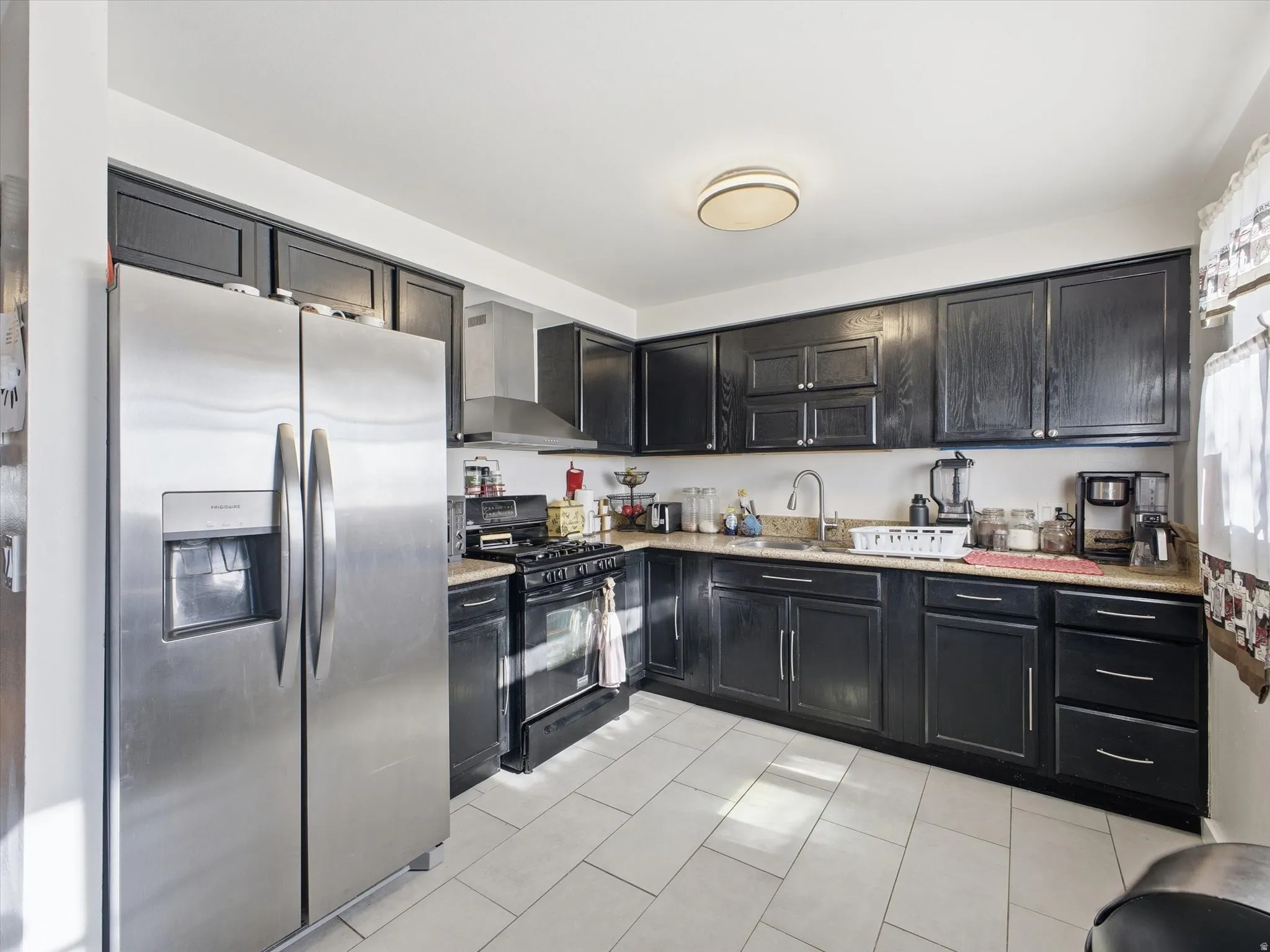 Kitchen featuring stainless steel fridge with ice dispenser, black gas range, dark cabinets, light tile patterned floors.