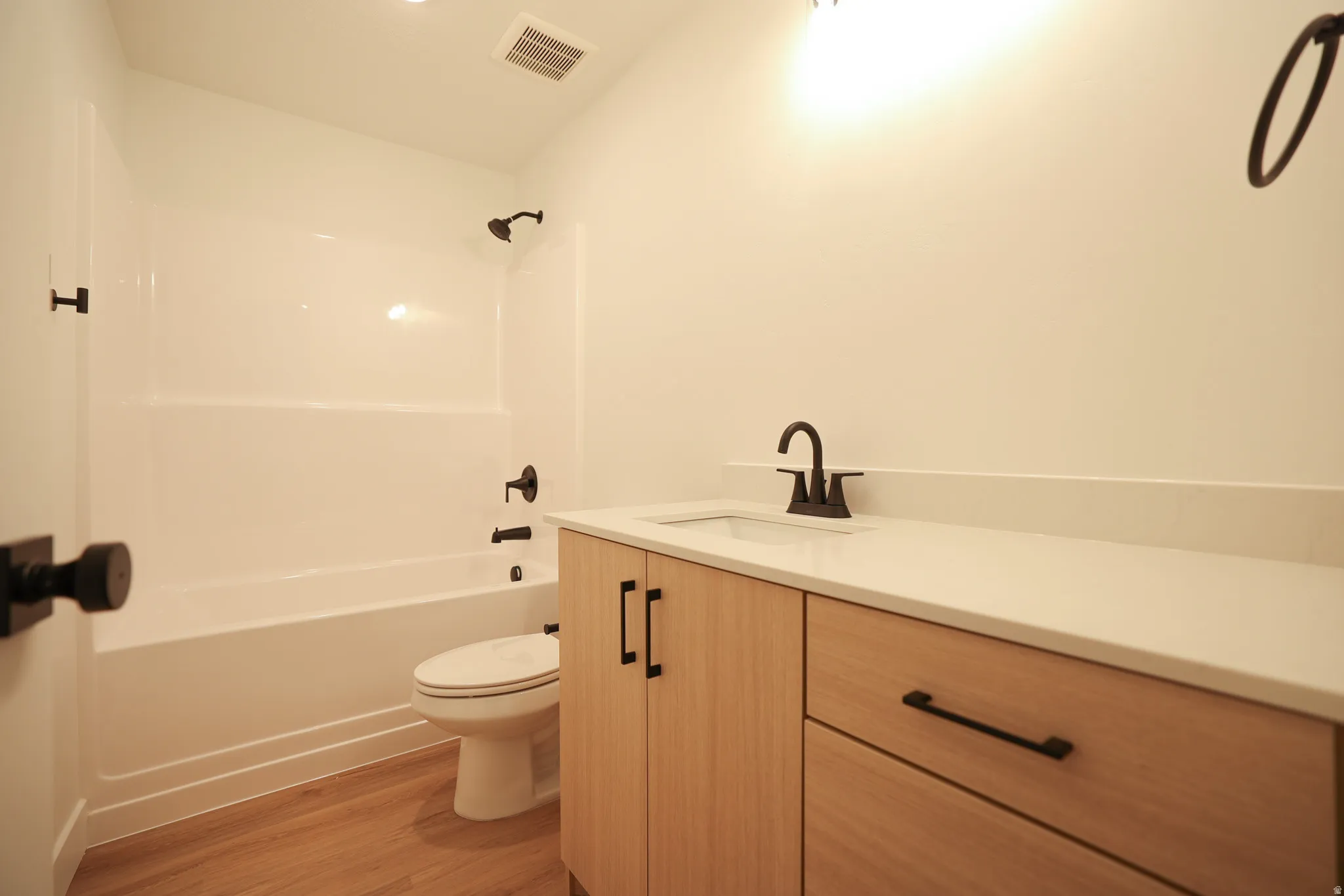 White tub/shower combination, light wood-finish vanity with built-in sink, stone-finish countertop, and wood-finish flooring