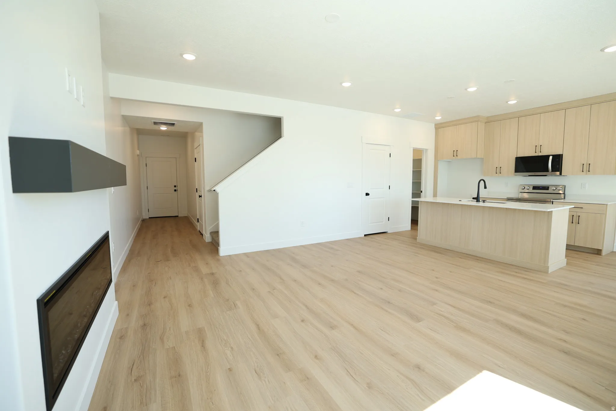 Open-concept living space featuring wood-finish flooring, a linear fireplace with a mantel, and a kitchen with light-toned cabinetry and a central island with an integrated sink