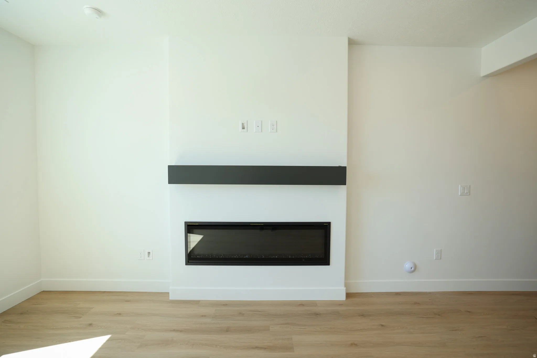 Built-in linear fireplace with a dark mantle, light wood-finish flooring, and crisp white walls