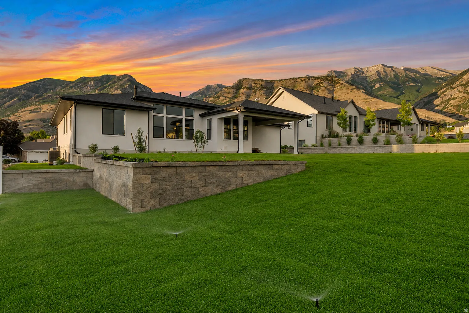 Expansive green lawn with visible irrigation system, retaining walls featuring stacked stone, and a modern stucco exterior with dark-framed windows