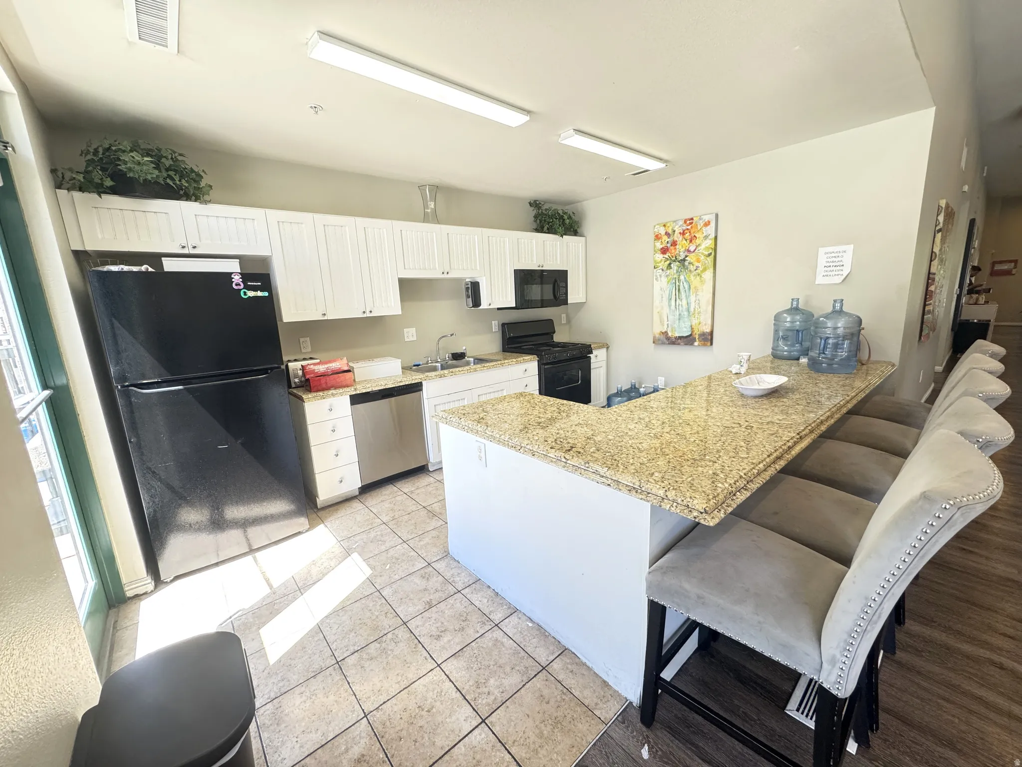 Club house itchen with a peninsula, black appliances, a kitchen bar, white cabinetry, and light tile patterned flooring