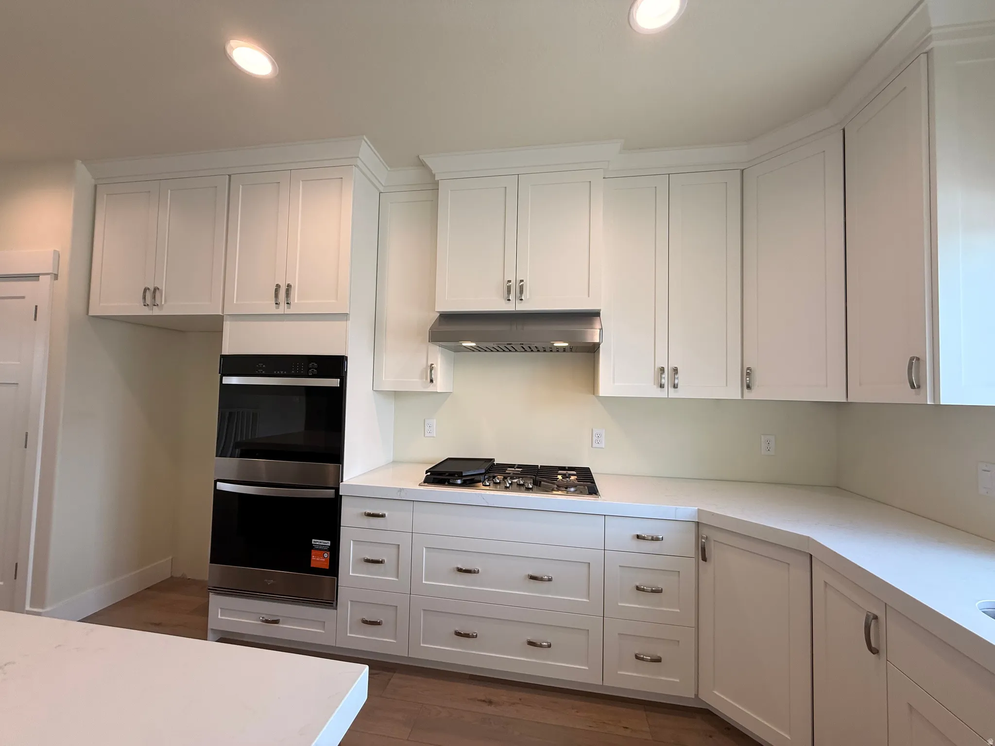 White shaker cabinetry, stainless steel double wall ovens, built-in gas cooktop with coordinating vent hood, wood-finish flooring, and light-toned countertops