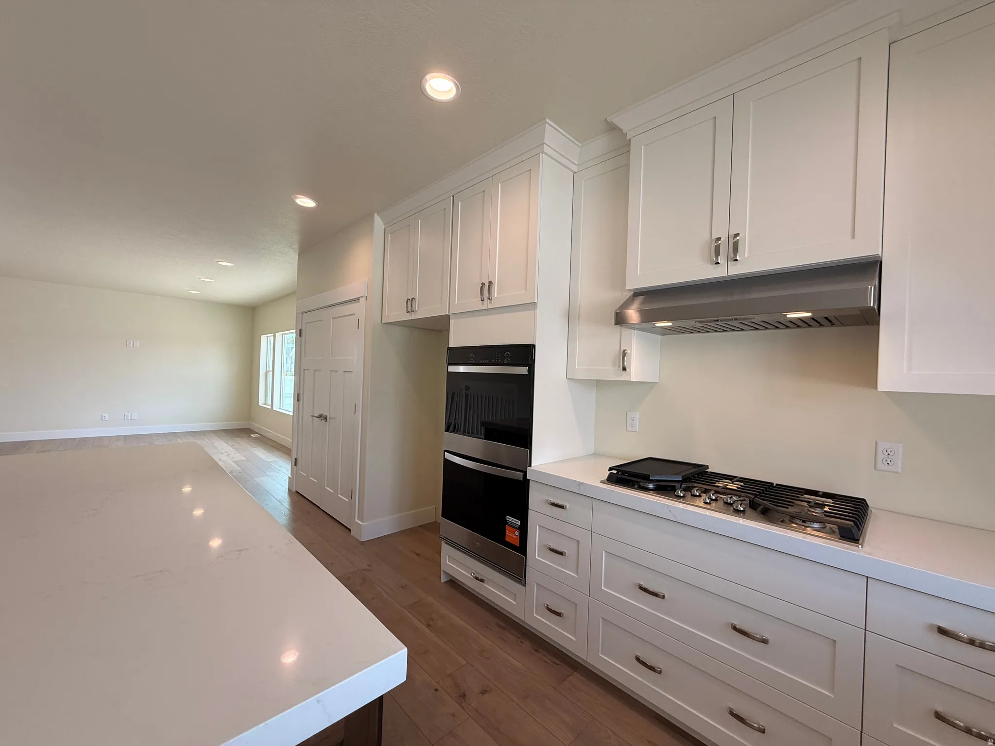 White shaker cabinetry, built-in double ovens, stainless steel range hood, built-in gas cooktop, and wood-finish flooring