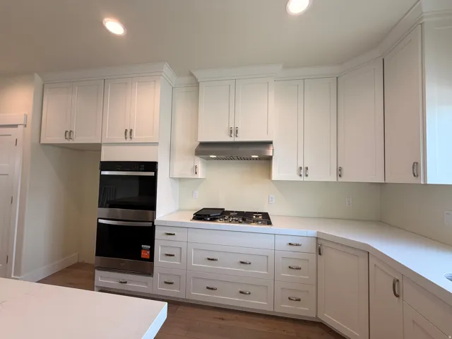 White shaker cabinetry, stainless steel double wall ovens, built-in gas cooktop with coordinating vent hood, wood-finish flooring, and light-toned countertops