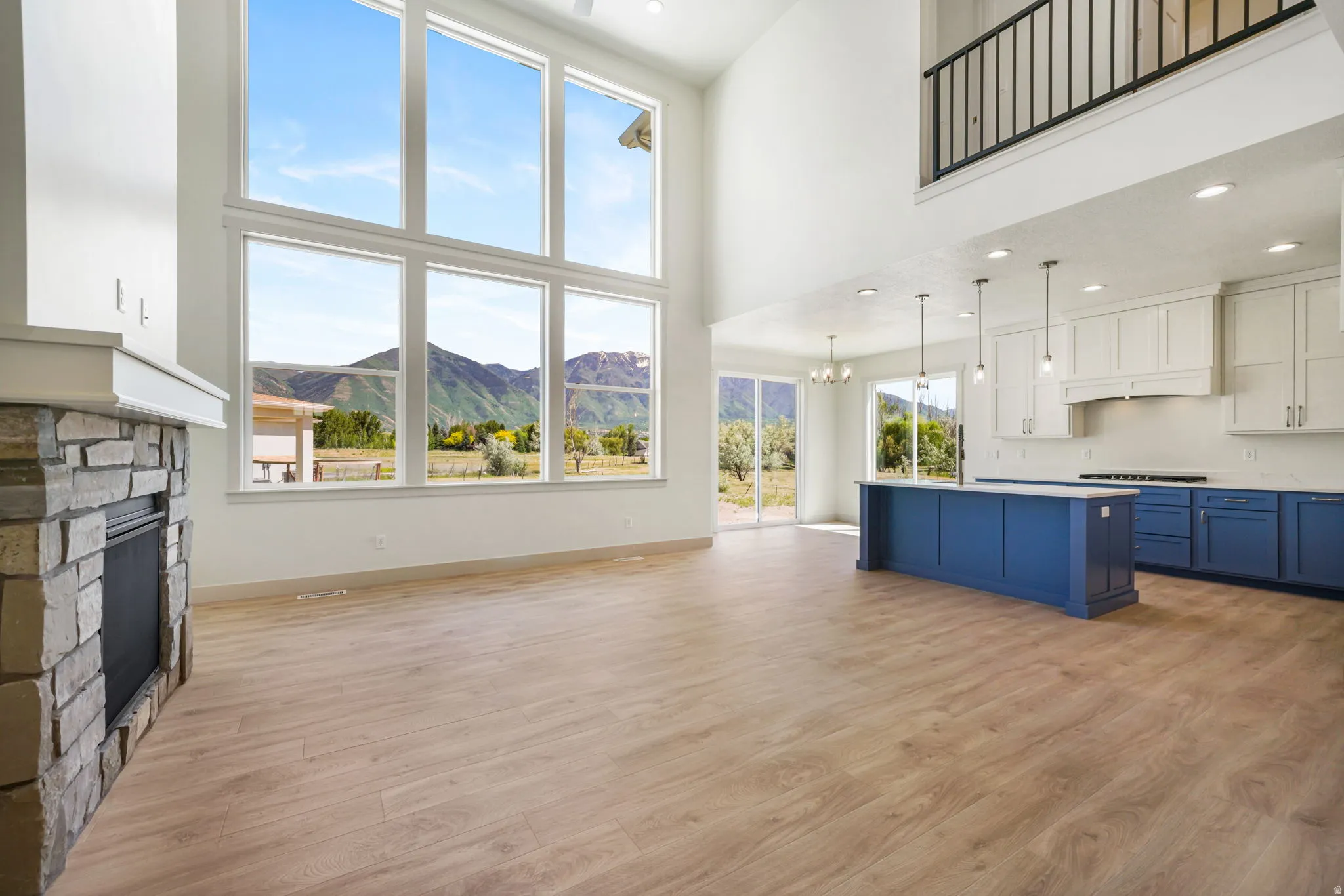 Two-story living area featuring a stone-clad fireplace, expansive windows, wood-finish flooring, and a kitchen with white upper and blue lower cabinetry