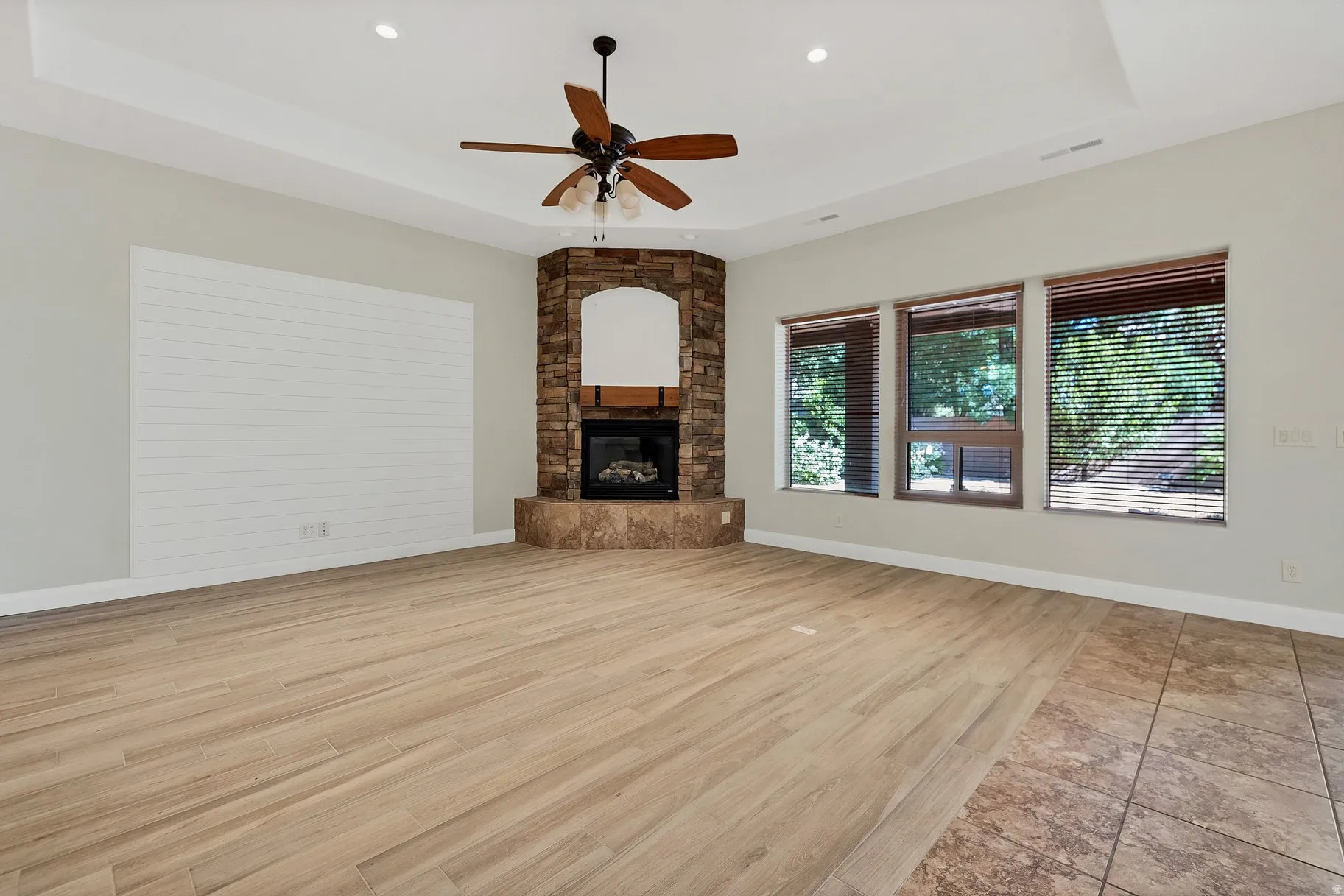 Unfurnished living room featuring a raised ceiling, light wood-style flooring, a ceiling fan, a stone fireplace, and recessed lighting