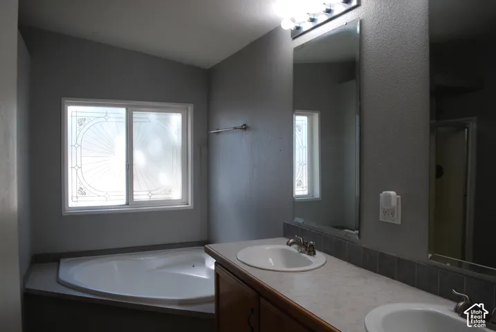 Corner soaking tub with decorative frosted window, dual vanity with integrated sinks, wood-finish cabinetry, and full-width mirror