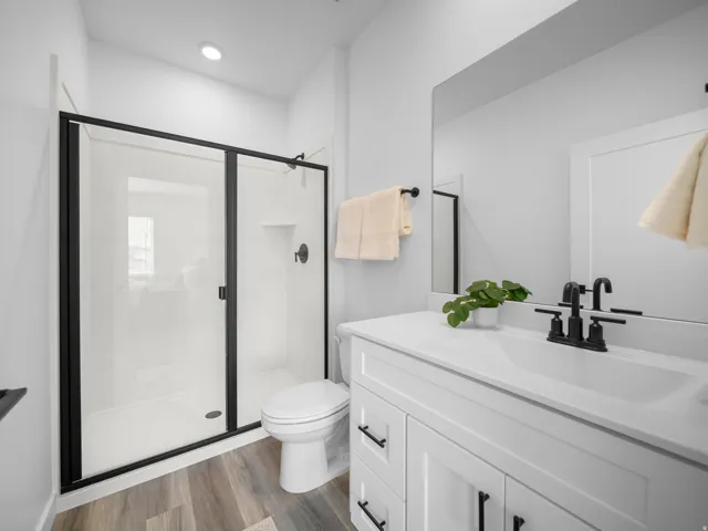 Walk-in shower with black-framed glass enclosure, white vanity with integrated sink and matte black faucet, full-width mirror, and wood-finish flooring