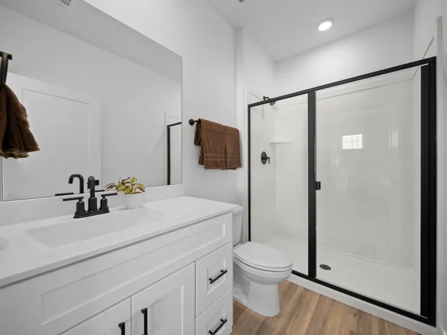 White vanity with integrated sink, black hardware, wood-finish flooring, and a walk-in shower with black-framed glass enclosure