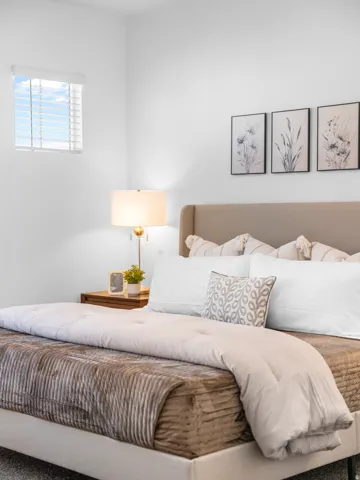 White walls, window with horizontal blinds, wood-finish nightstand, upholstered headboard, and a soft gray carpet