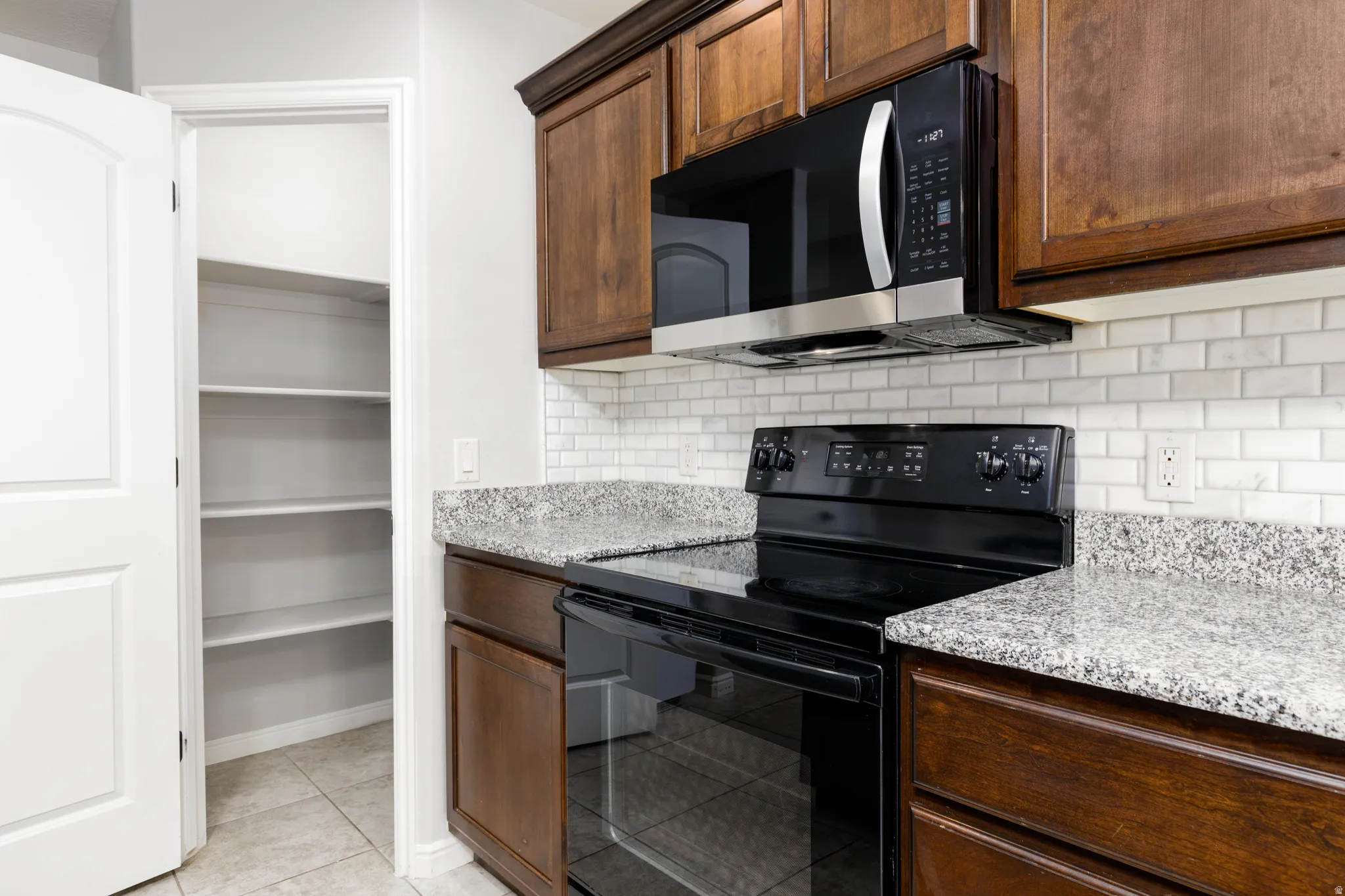 Kitchen with electric range, grantie counters, subway stone tile, light tile floors, dark wood finish cabinets and pantry