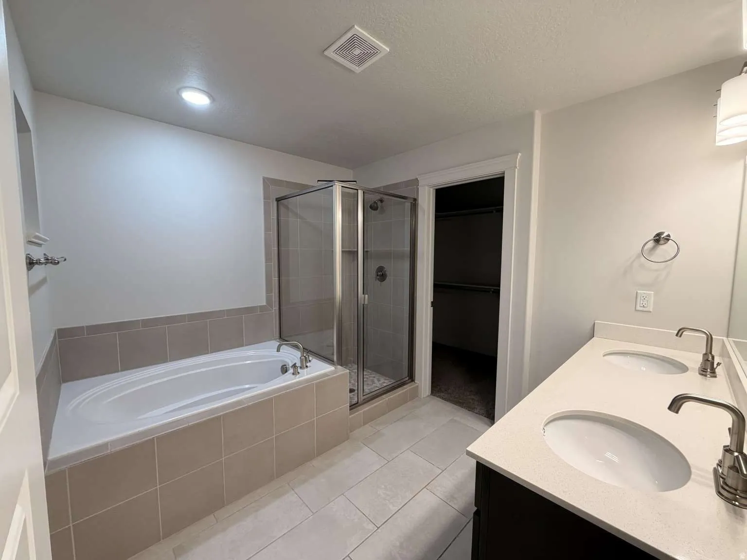 Bathroom featuring a dual-sink vanity with light-toned countertops, a built-in soaking tub with tiled surround, and a glass-enclosed shower