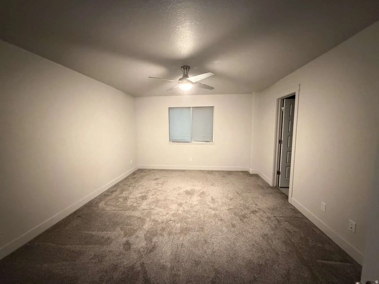 Spacious interior room featuring light-colored walls, textured carpet flooring, and a ceiling fan with integrated lighting