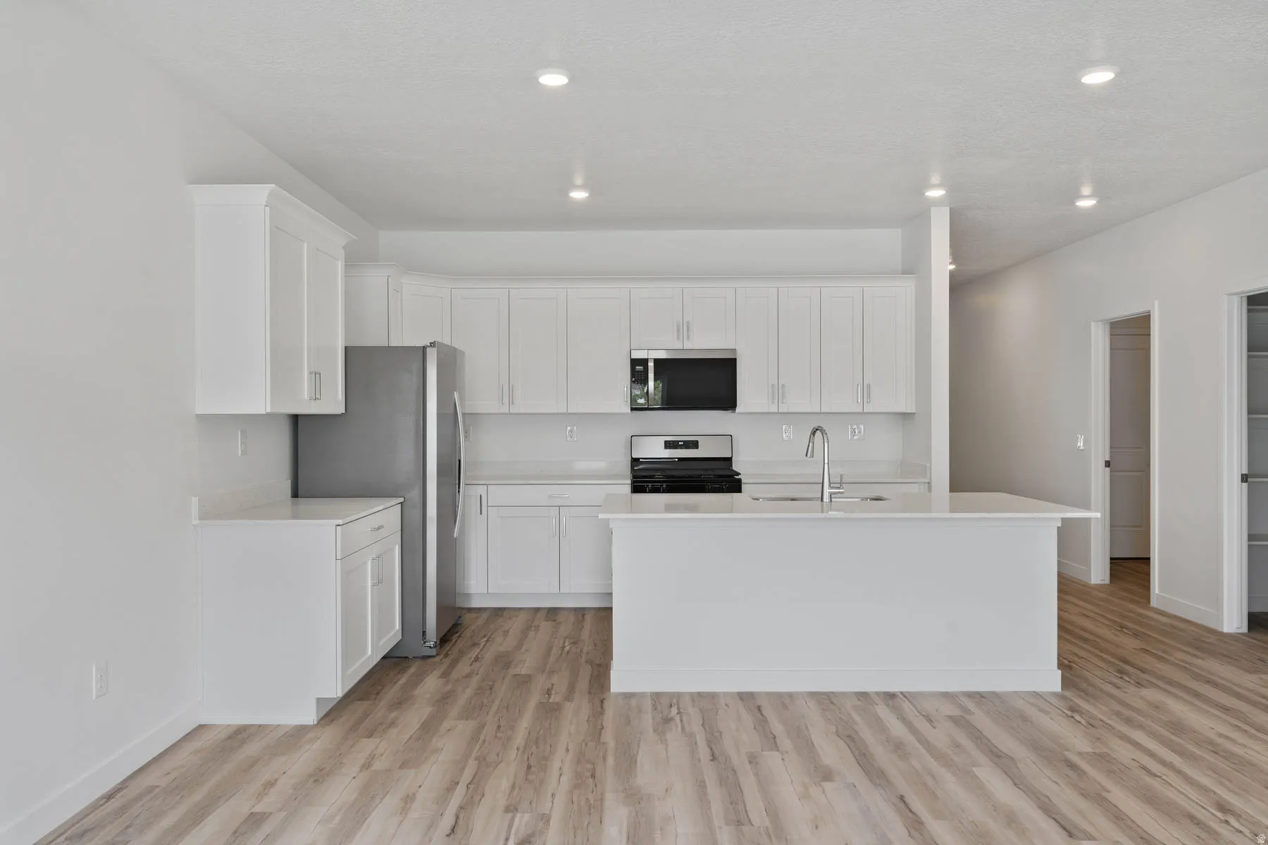 Contemporary kitchen featuring white shaker cabinetry, stainless steel appliances, light-toned countertops, a central island with sink, and wood-finish flooring