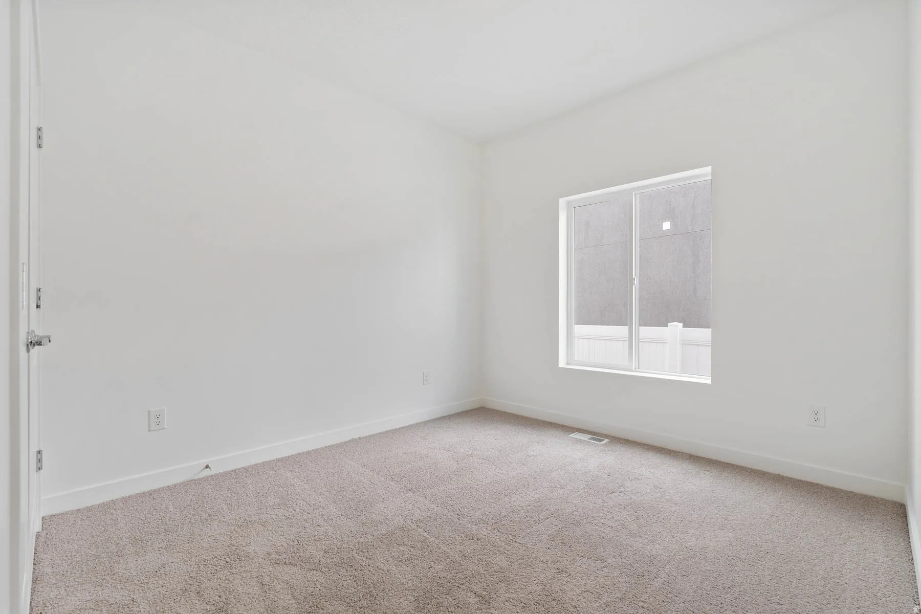 Bright interior room featuring a large window, neutral carpeting, white baseboards, and a solid core door with brushed nickel hardware