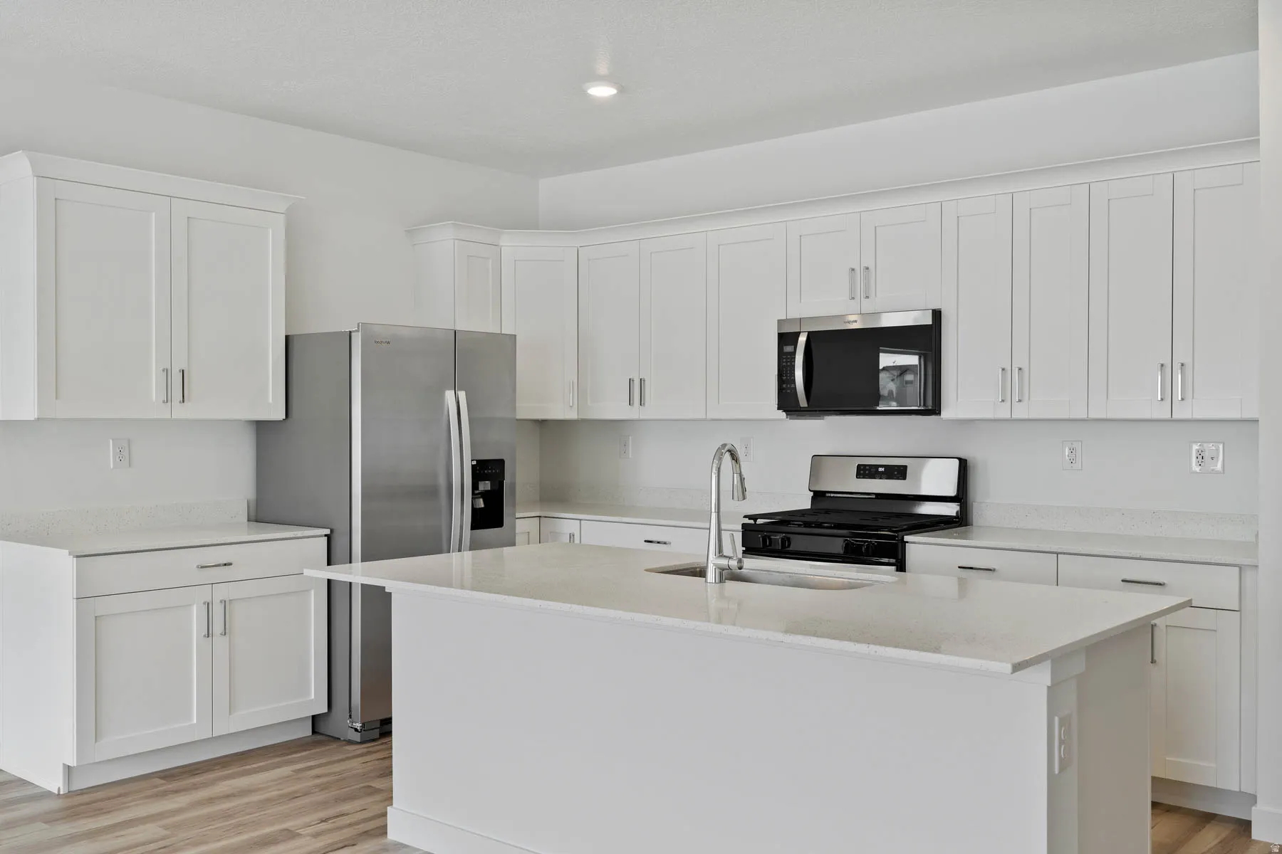 Kitchen featuring white shaker cabinetry, stainless steel appliances, light-toned countertops, a central island with an undermount sink, and wood-finish flooring