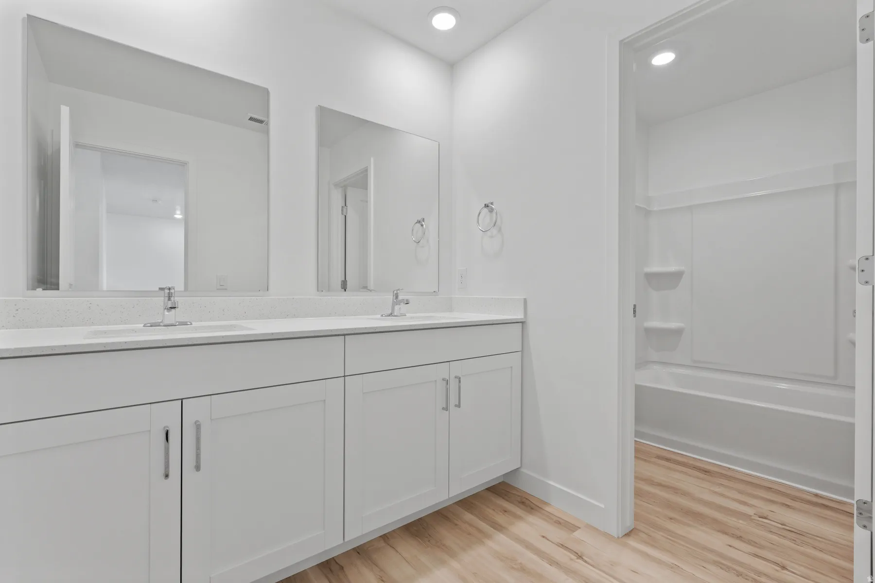 Dual sink vanity with white cabinetry, light-toned wood-finish flooring, recessed lighting, white solid surface countertops, and a white tub/shower combination