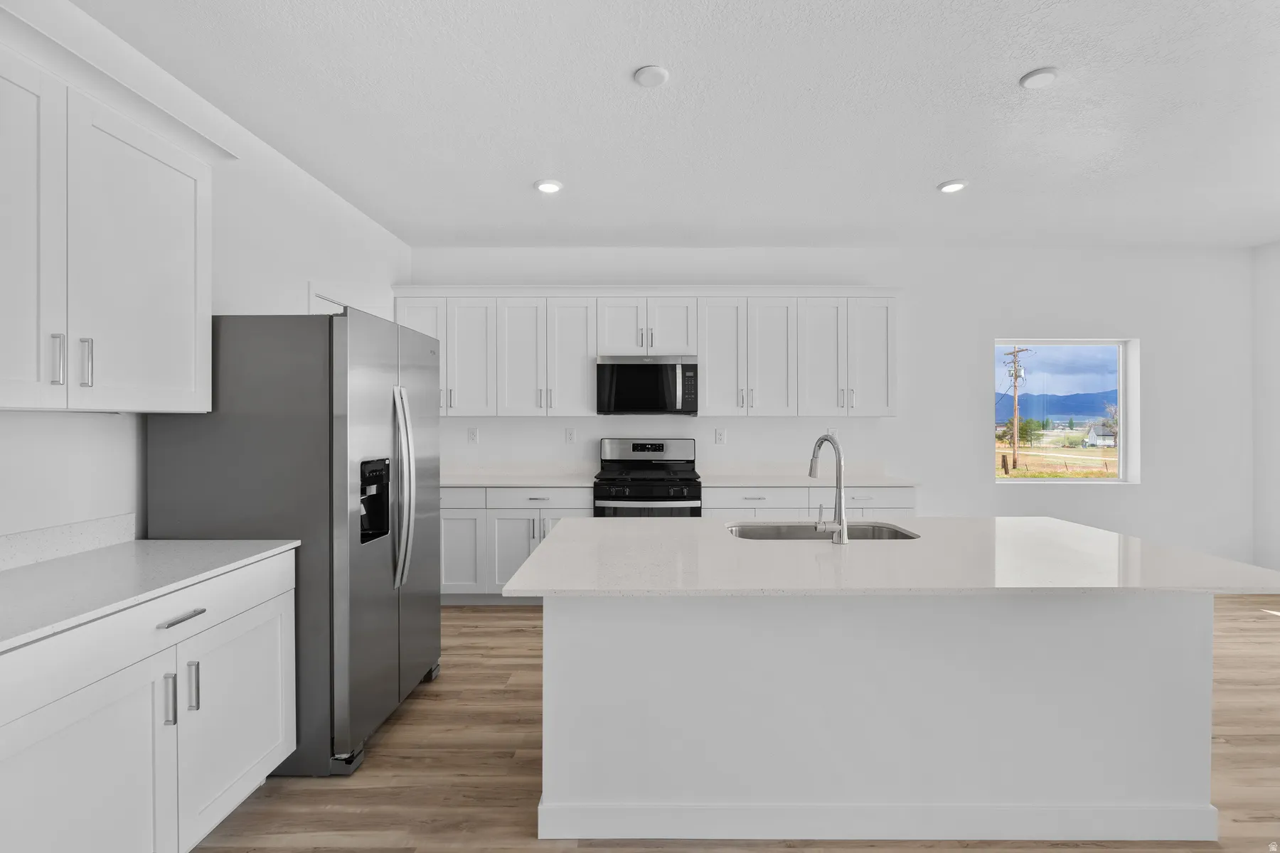 Modern kitchen featuring white cabinetry, light-toned countertops, stainless steel appliances, a kitchen island with an integrated sink, and wood-finish flooring