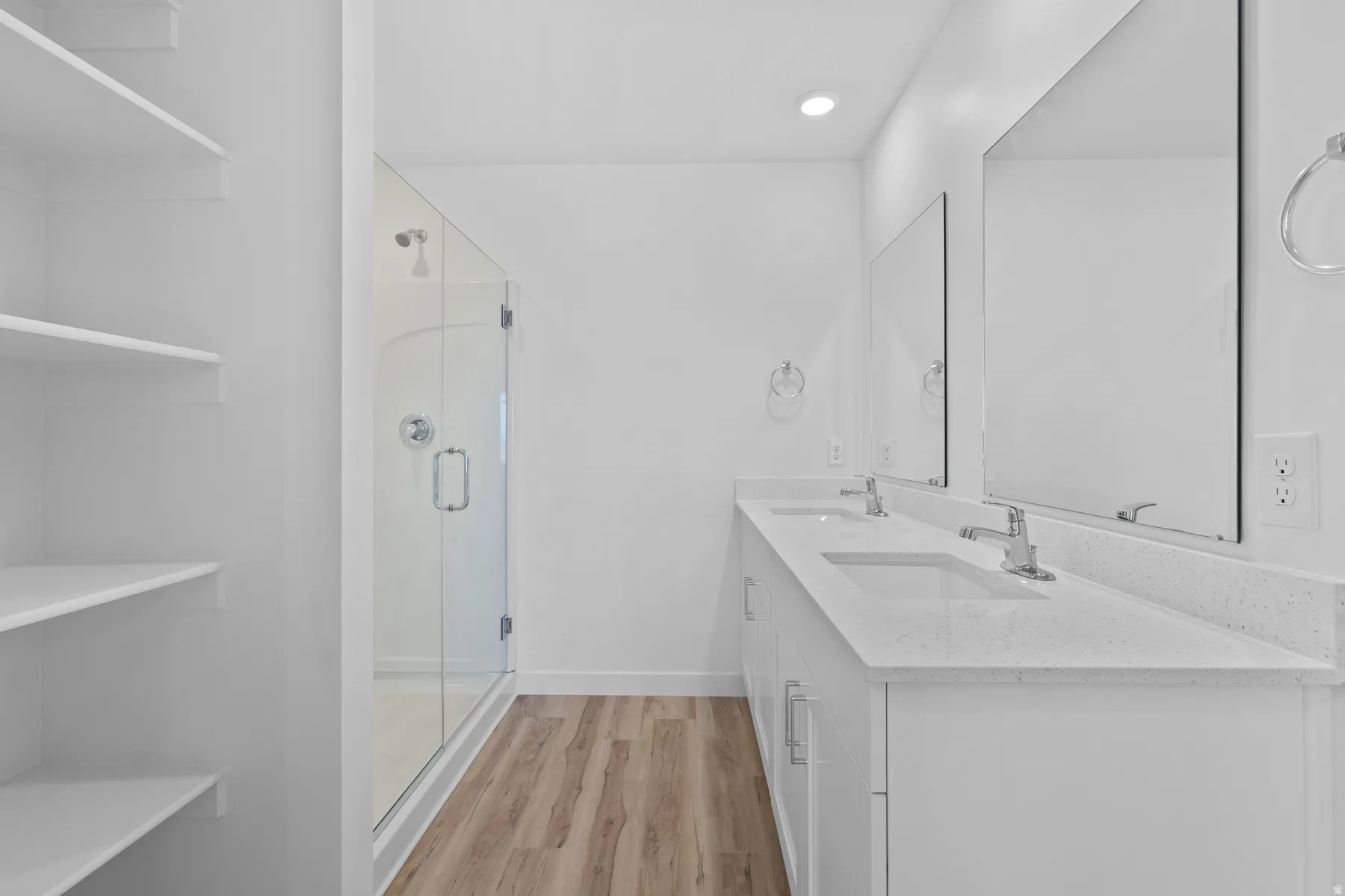 Bathroom featuring a dual vanity with white cabinetry and integrated sinks, a glass-enclosed shower, wood-finish flooring, and built-in shelving