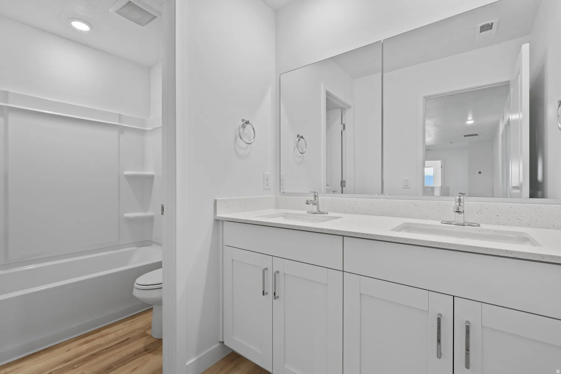 Bathroom featuring a dual vanity with white cabinetry and light-toned countertops, a large wall-mounted mirror, and wood-finish flooring