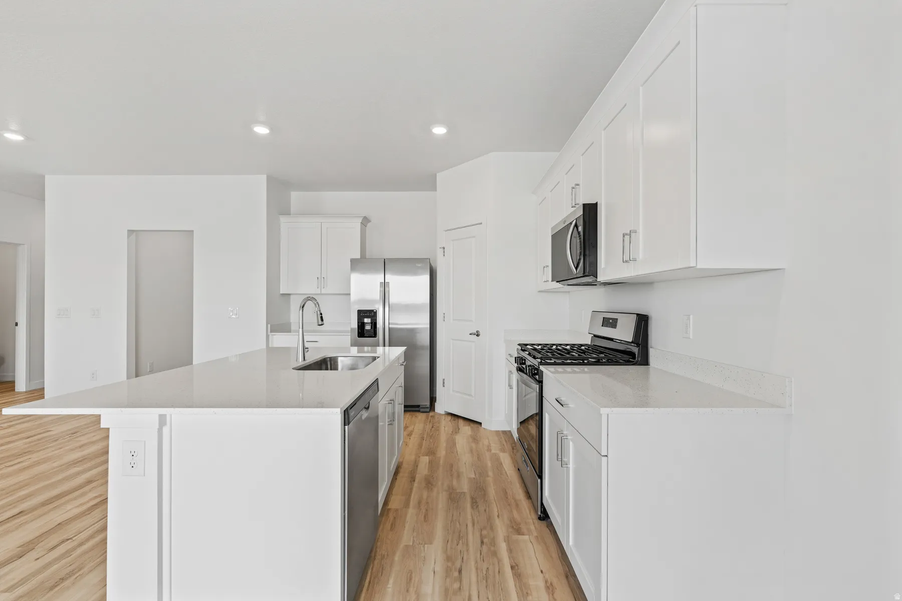 Spacious kitchen featuring an expansive island with an undermount sink, stainless steel appliances, white cabinetry, light countertops, and wood-finish flooring