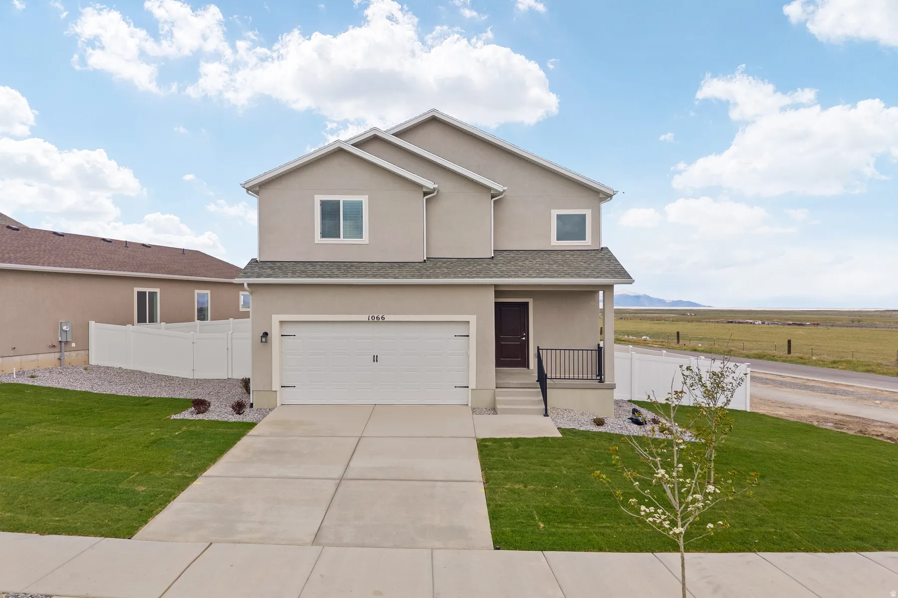 Contemporary two-story residence featuring a neutral stucco exterior, a two-car garage, a covered front porch with dark railings, a concrete driveway, and a manicured lawn