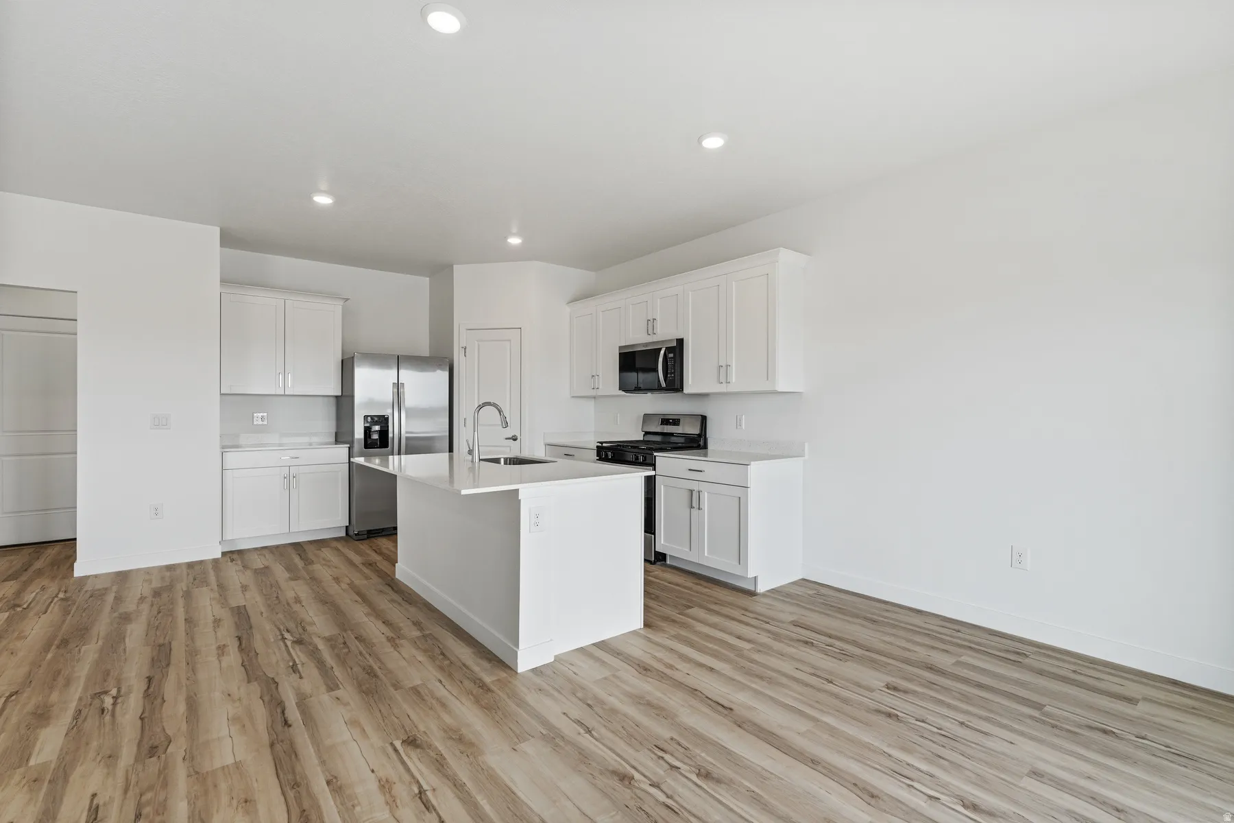 Modern kitchen featuring a central island with an undermount sink, white cabinetry, integrated stainless steel appliances, recessed lighting, and wood-finish flooring
