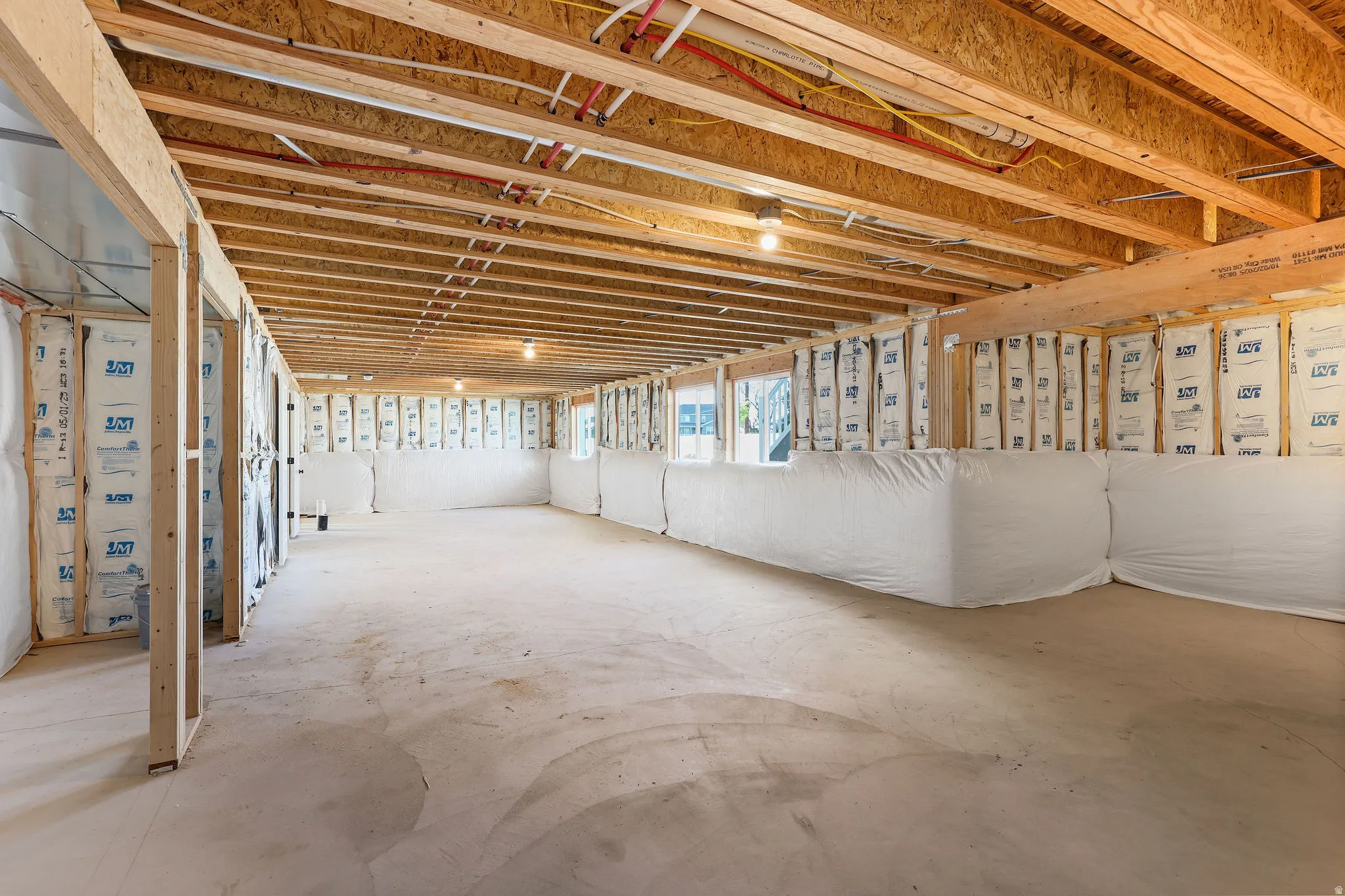Unfinished basement space featuring exposed wood joists and subflooring, framed walls with insulation, concrete flooring, and multiple light fixtures