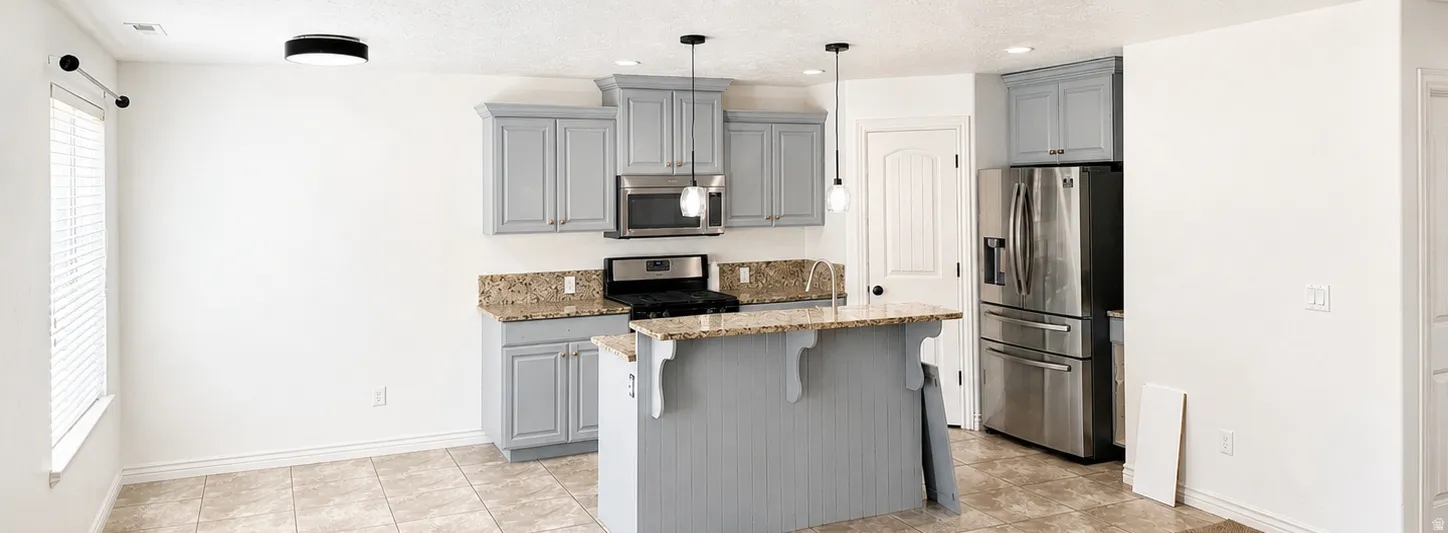 Kitchen featuring a central island with natural stone countertop, light-toned cabinetry, stainless steel appliances, tiled flooring, and modern pendant lighting