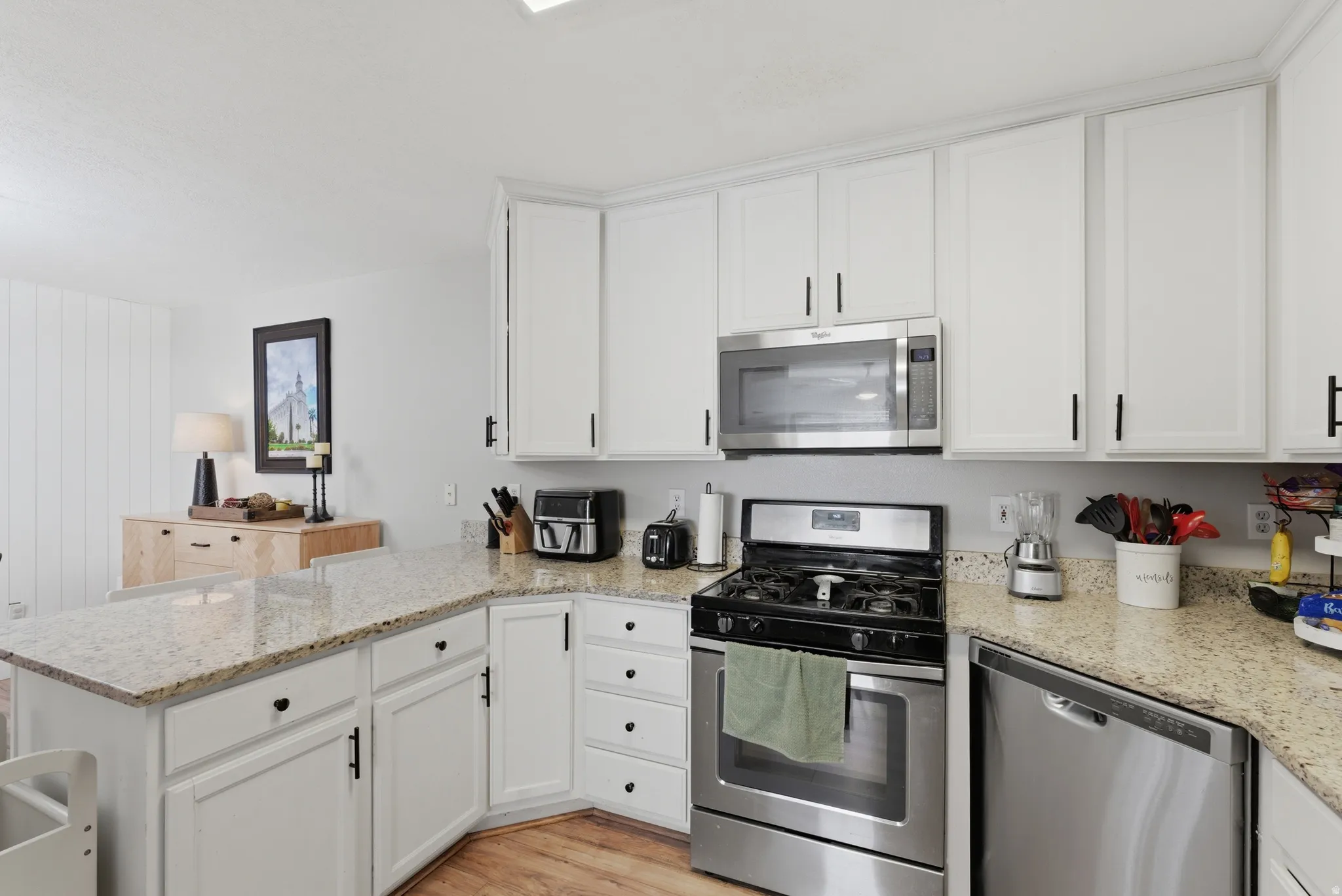 Kitchen with stainless steel appliances, a peninsula, white cabinets, light wood-type flooring, and light stone countertops