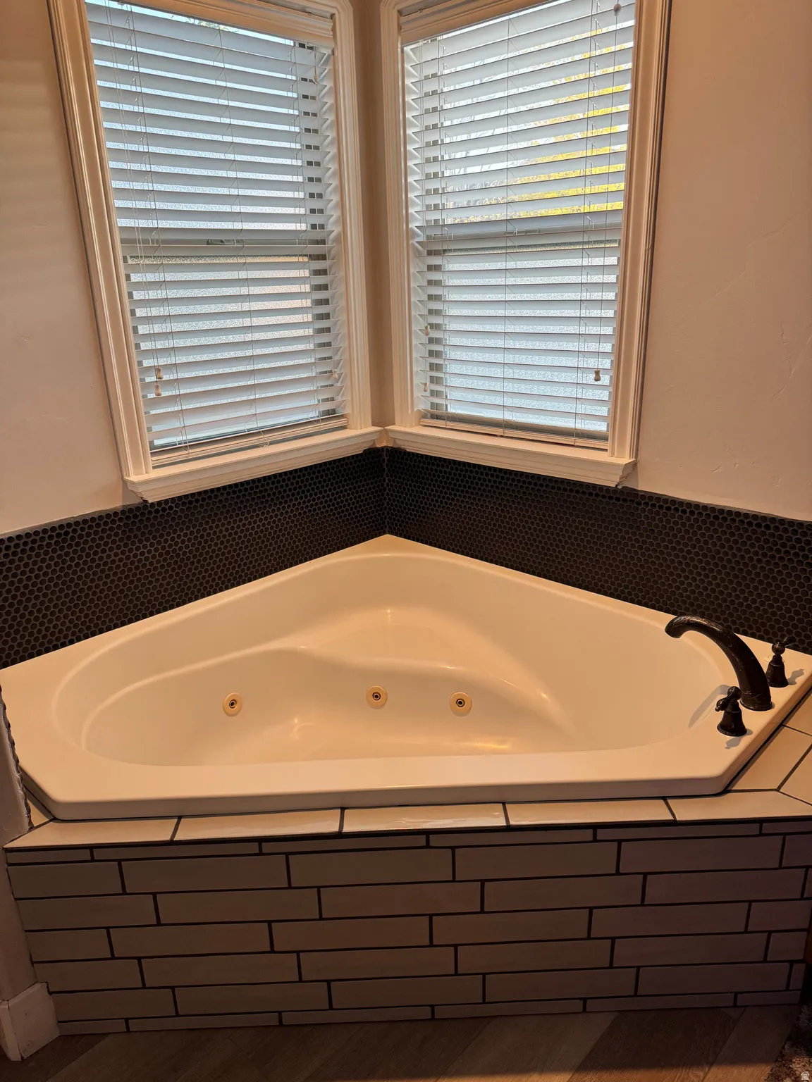 Corner jetted tub featuring a bronze-finish faucet, black penny tile backsplash, and light wood-tone subway tile surround