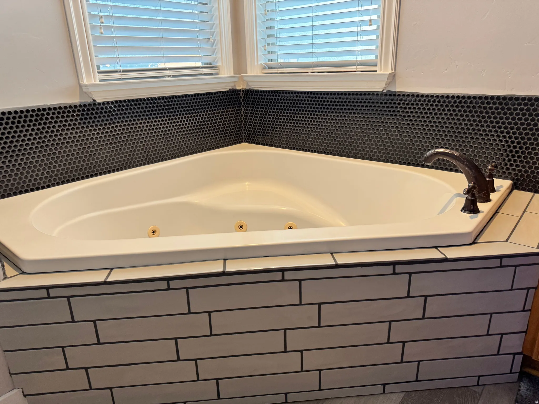 Corner jetted tub featuring a bronze-finish faucet, black penny tile backsplash, and light subway tile surround