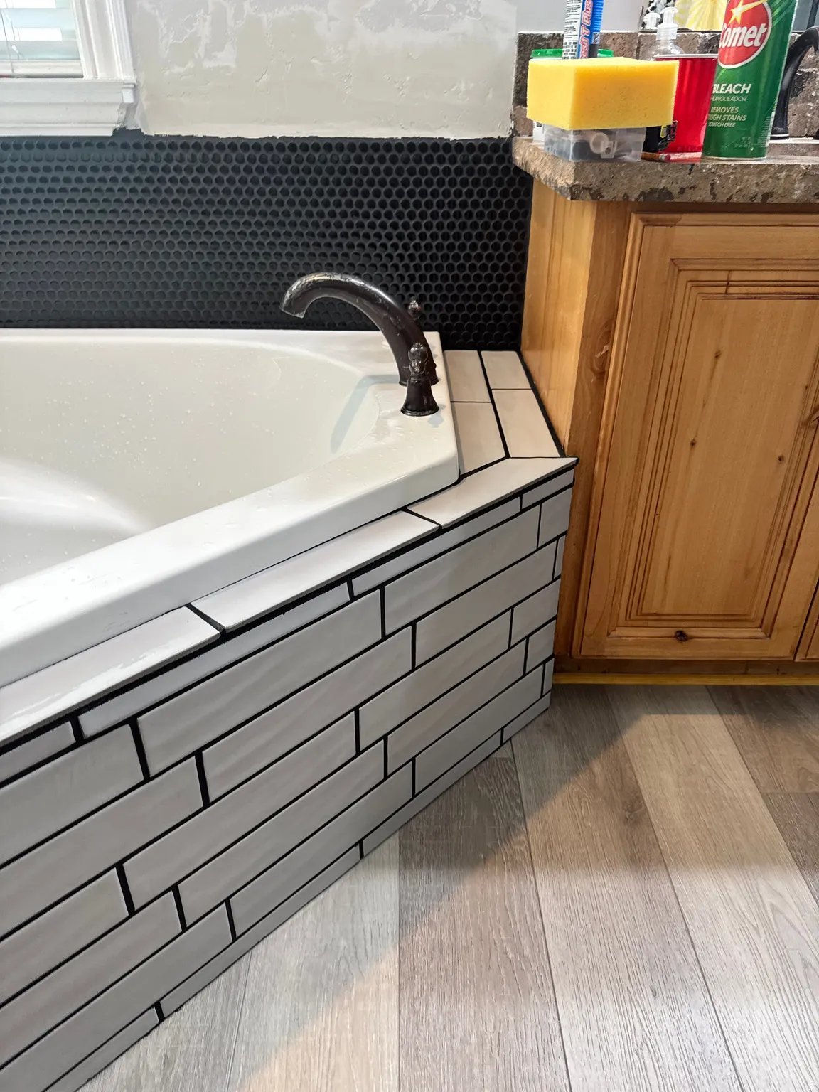Soaking tub featuring a bronze-finish faucet, black penny tile backsplash, light wood-finish flooring, and a stone-look countertop on wooden cabinetry
