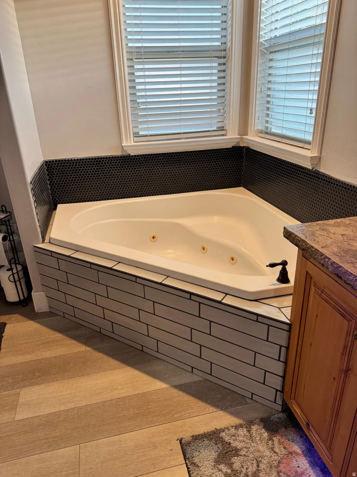 Corner jetted tub featuring a dark penny tile surround and wood-finish flooring