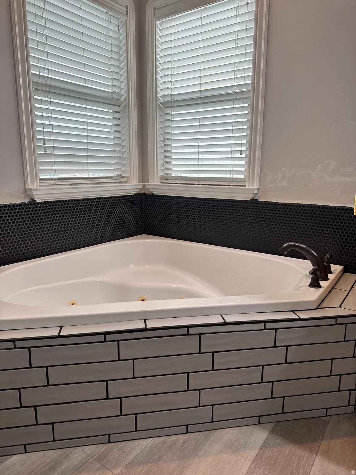 Corner jetted tub featuring a bronze-finish faucet, black penny tile surround, and horizontal stacked tile skirt