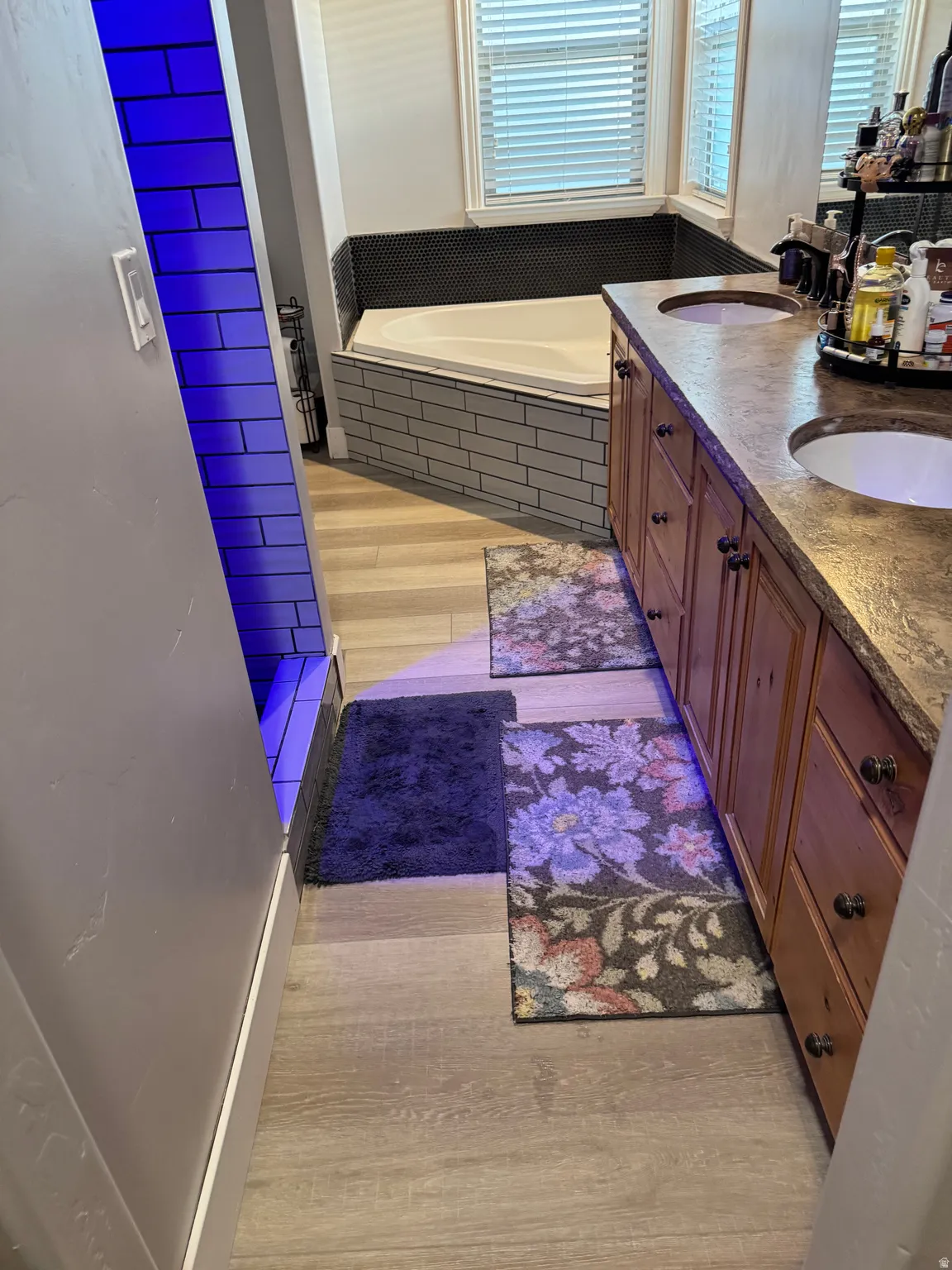 Bathroom featuring a built-in soaking tub with subway tile surround, dual vanity with undermount sinks and stone-finish countertop, and wood-finish flooring