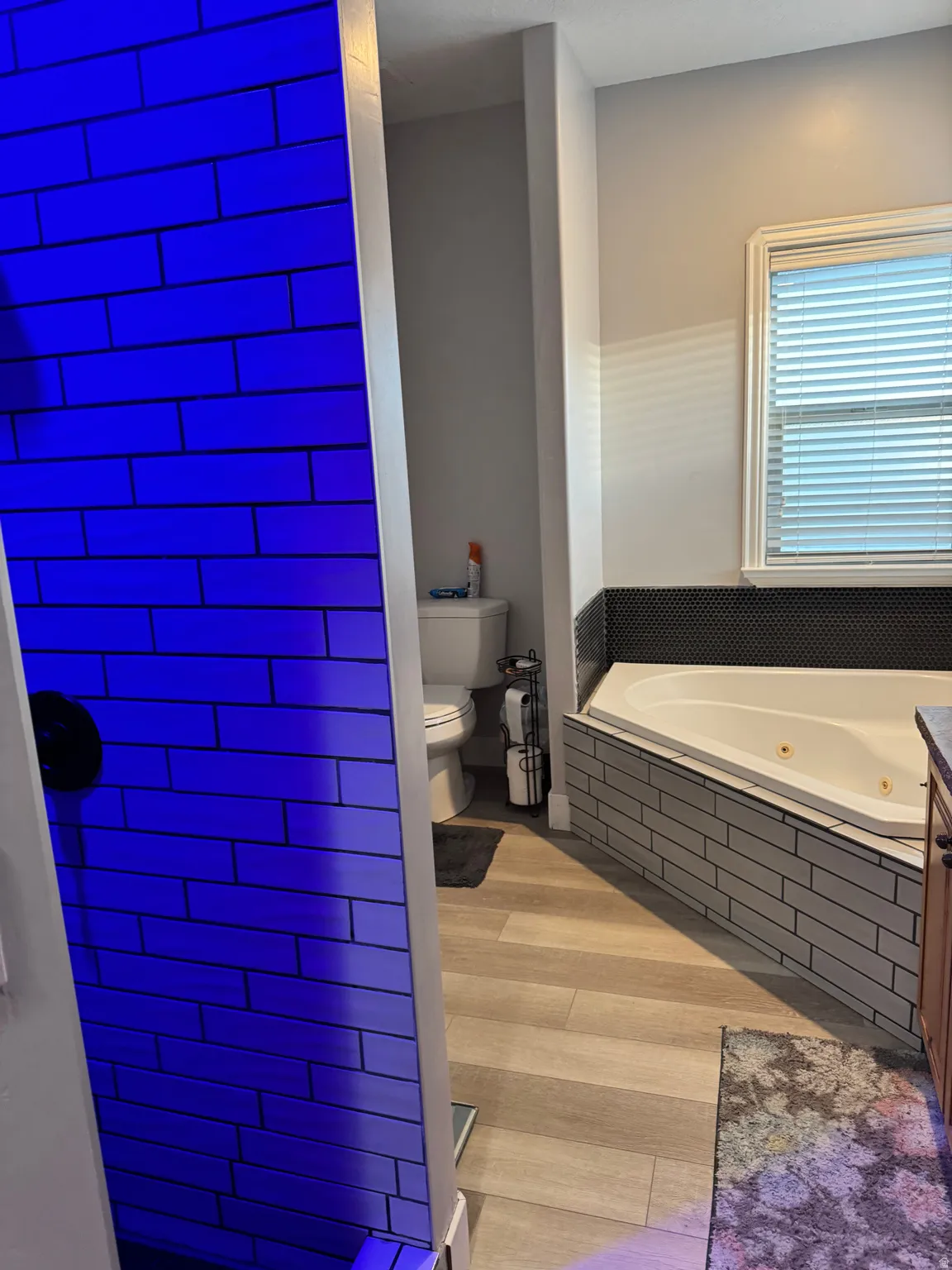 Bathroom featuring wood-finish flooring, a jetted corner tub with subway tile surround, a toilet, and a tiled shower stall with a dark blue light accent