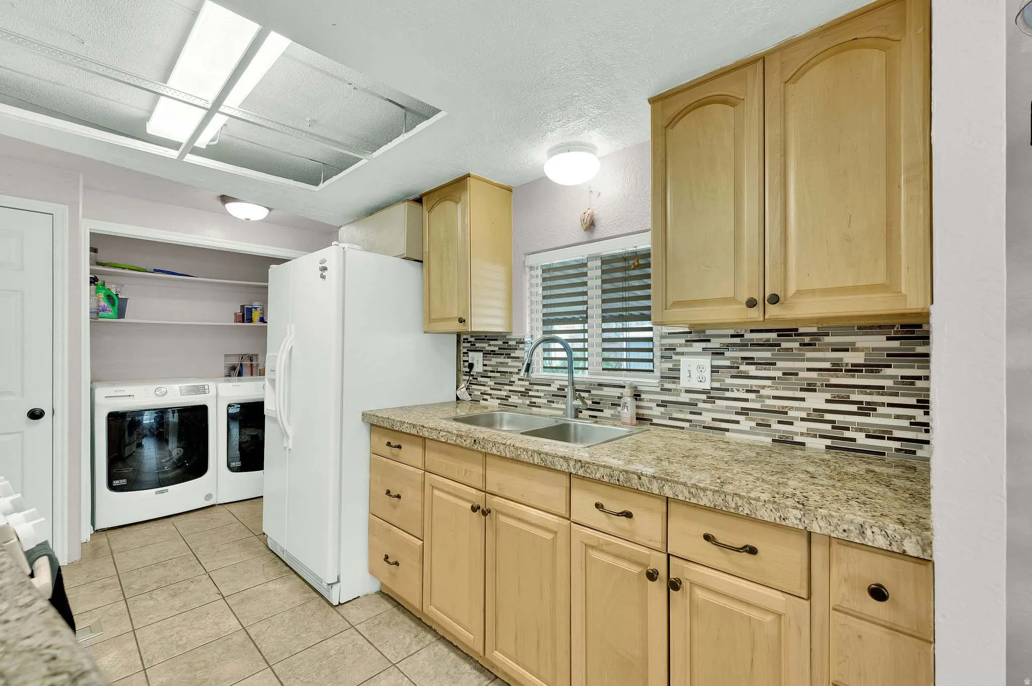 Kitchen featuring light wood finish cabinets, white fridge with ice dispenser, washer and clothes dryer, and light tile patterned floors