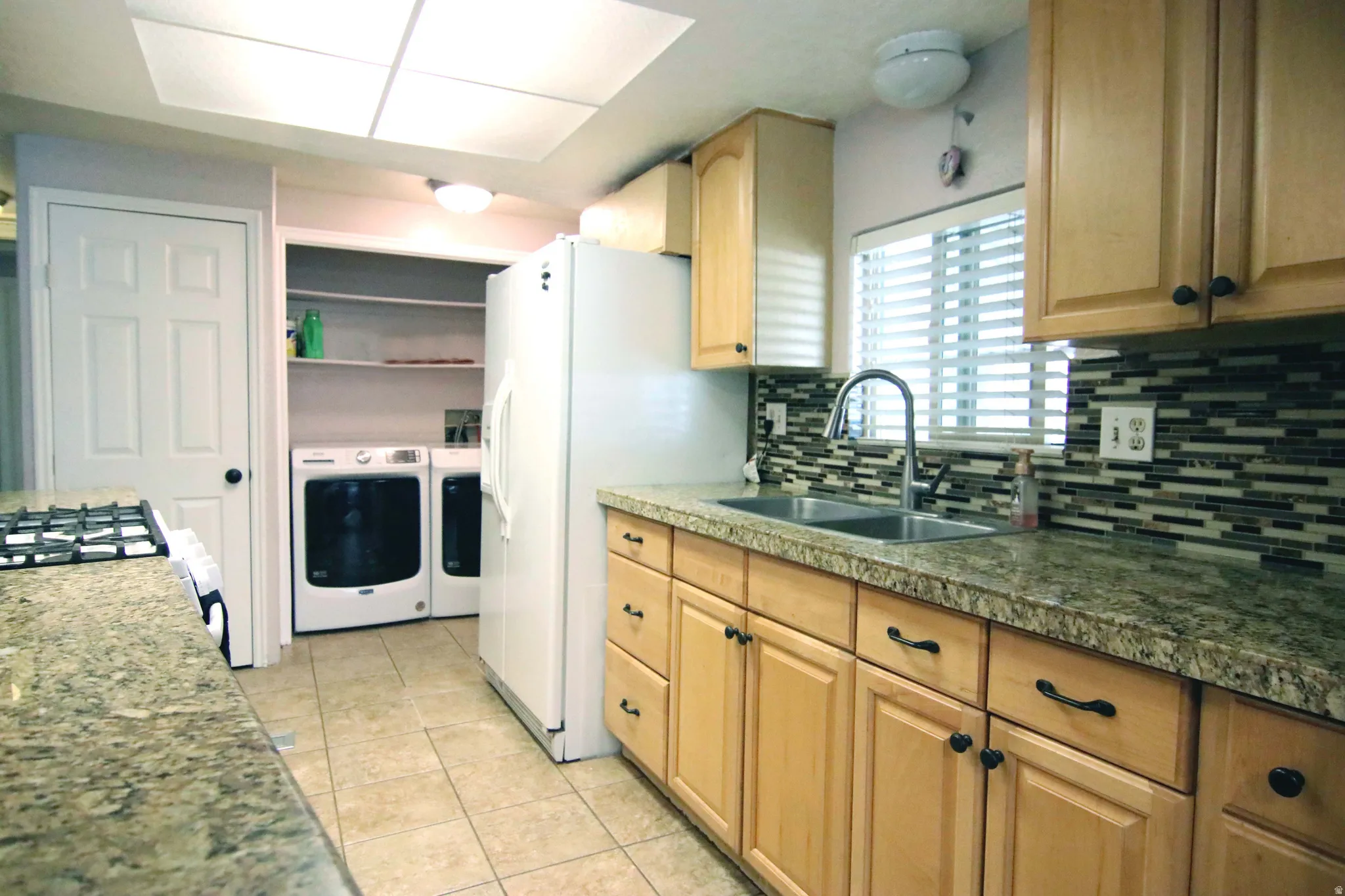 Kitchen with separate washer and dryer, light wood finish cabinetry, light tile patterned flooring, white refrigerator with ice dispenser, and gas stove