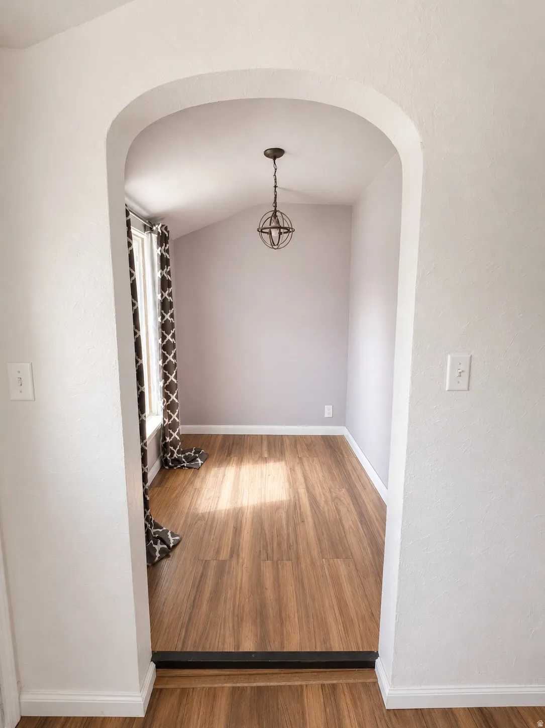 Archway entry to a room featuring wood-finish flooring, a contemporary spherical pendant light, and a window with patterned curtains