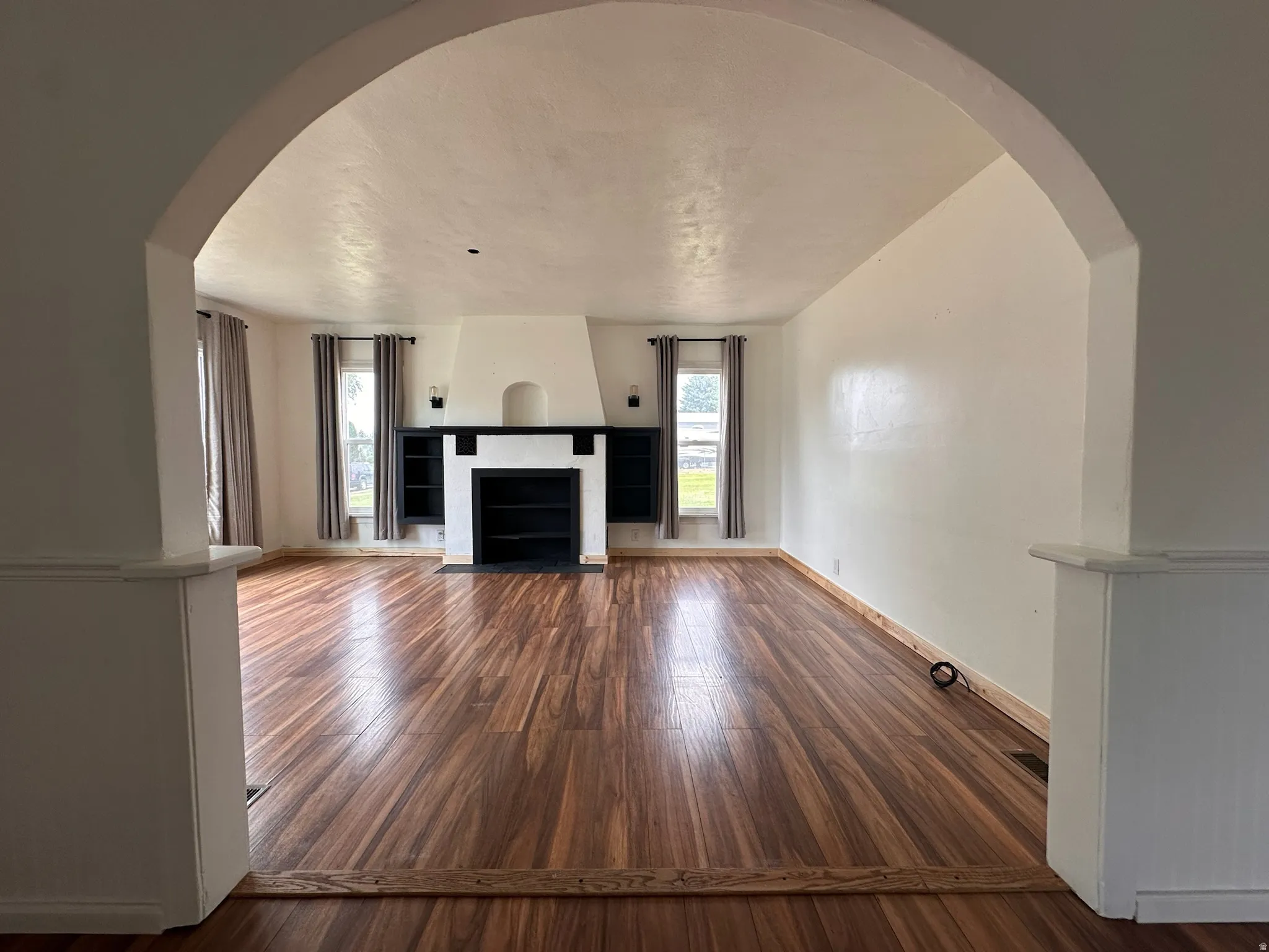 Archway entry to a room featuring wood-finish flooring, a central fireplace with a dark mantel, built-in shelving, and multiple windows with drapery