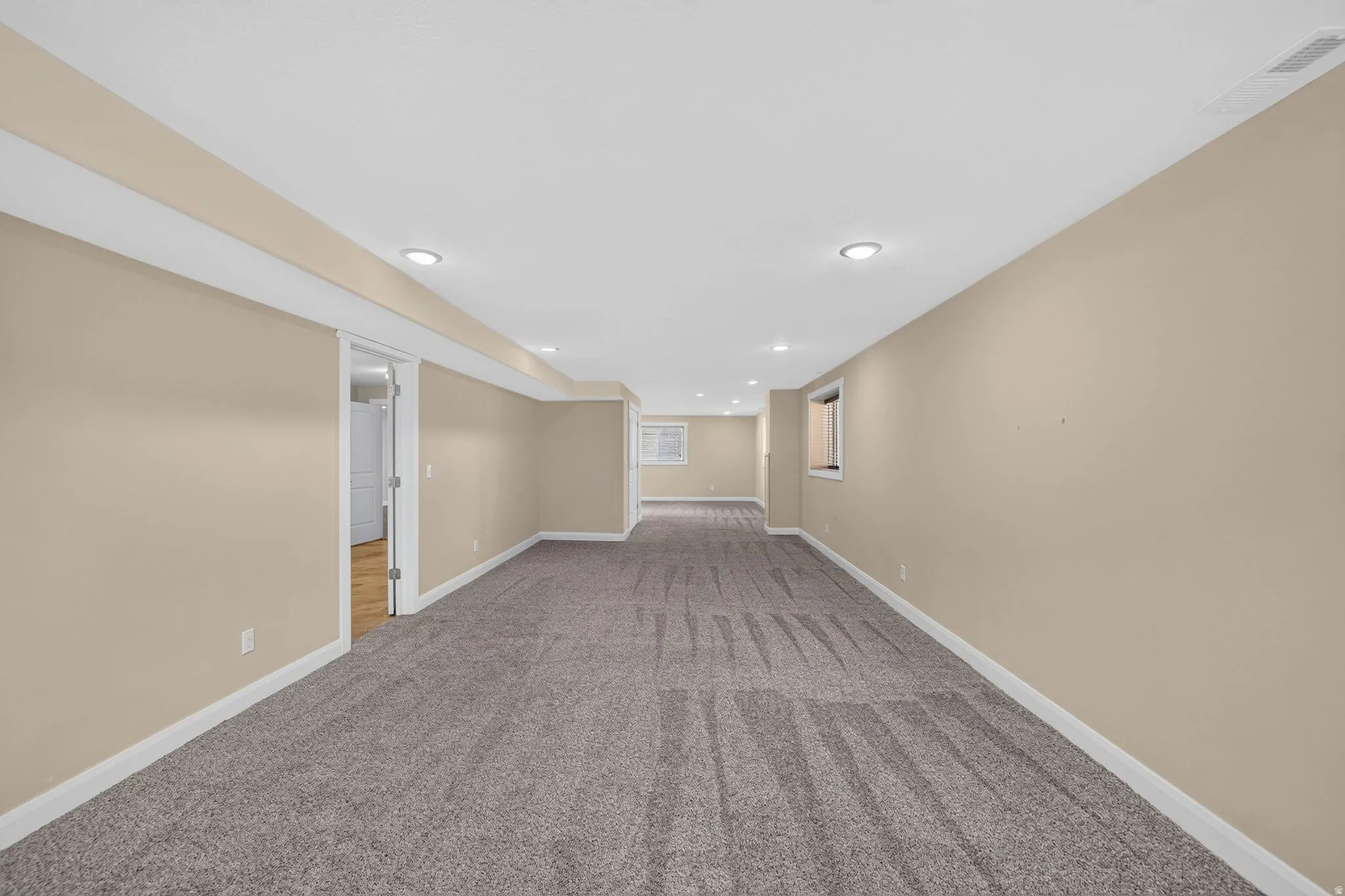 Spacious finished basement with neutral-toned carpeting, recessed lighting, light beige walls, white trim, and a doorway leading to a separate area with wood-finish flooring
