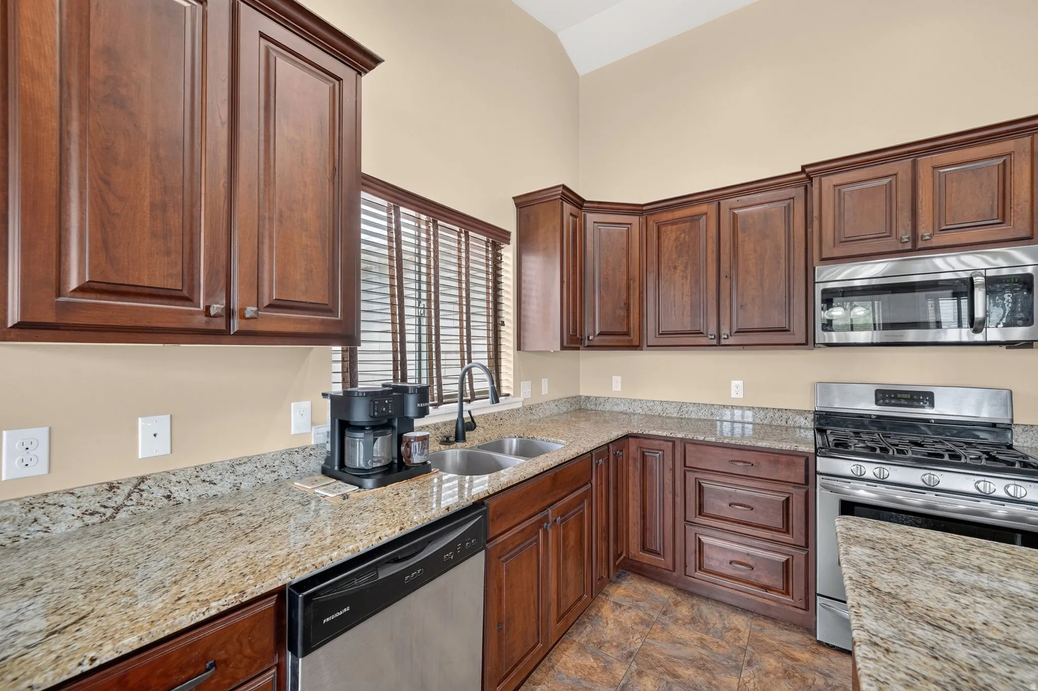 Kitchen featuring rich wood cabinetry, granite countertops, a stainless steel gas range, a double basin sink with a gooseneck faucet, and durable tile flooring