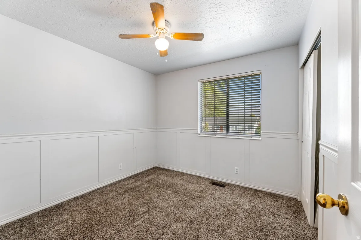 Empty room featuring dark carpet, a decorative wall, ceiling fan, a textured ceiling, and a wainscoted wall
