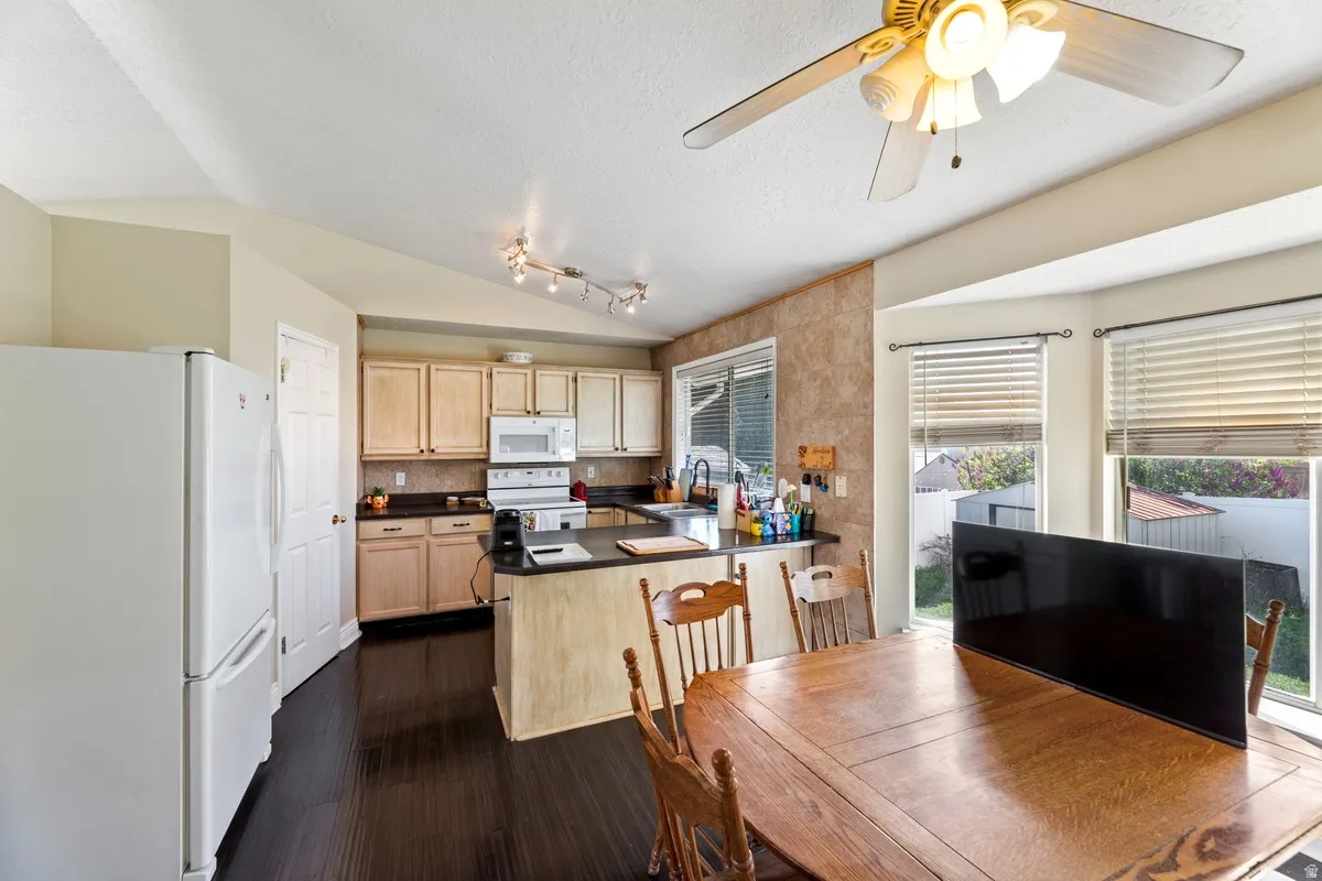 Kitchen featuring white appliances, a peninsula, dark countertops, dark wood-style flooring, and ceiling fan