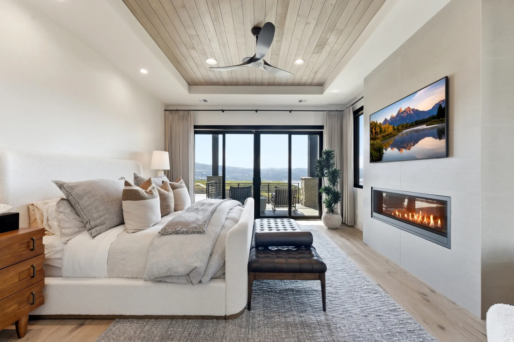 Bedroom featuring access to exterior, a wood tray ceiling, light wood finished floors, a glass covered fireplace, and a ceiling fan