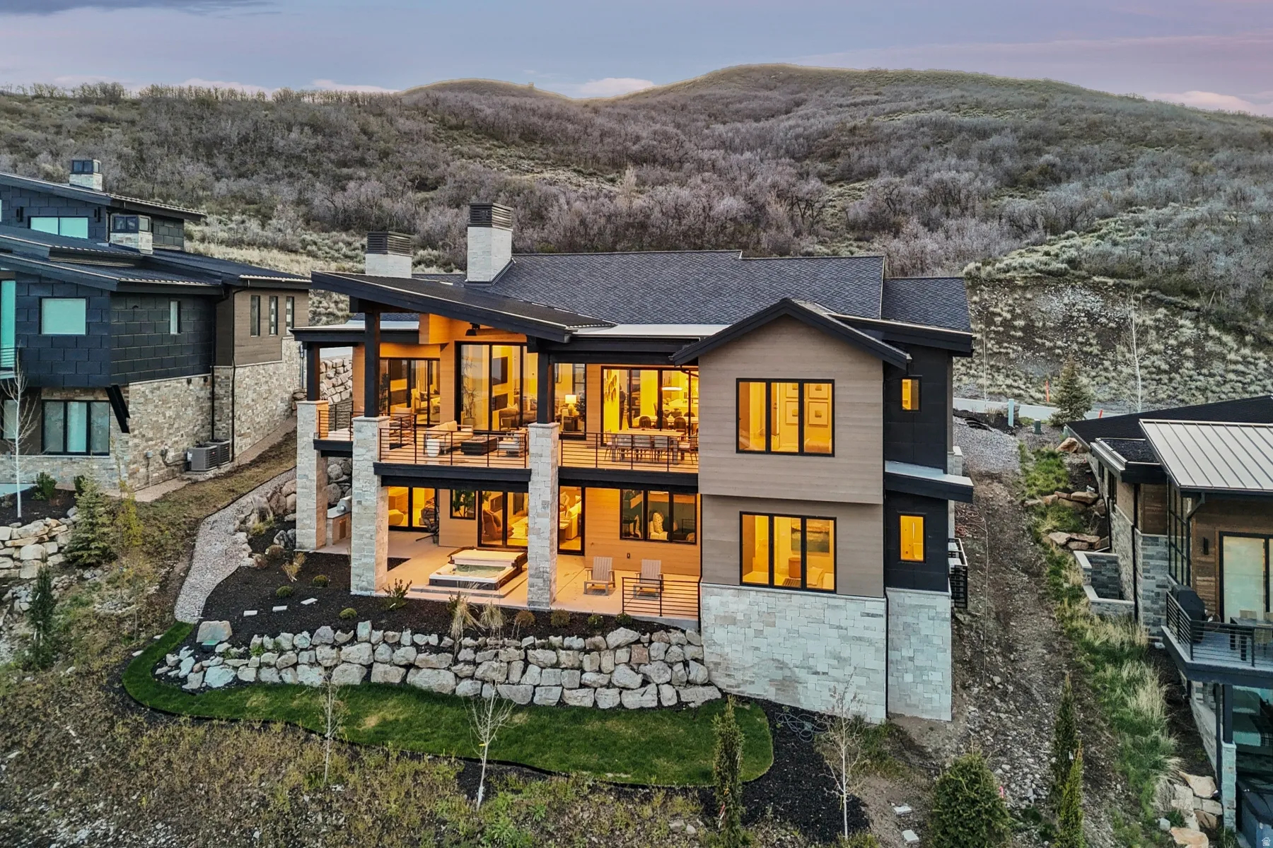 Back of house at dusk with stone siding, a chimney, a patio area, a wooded view, and a balcony