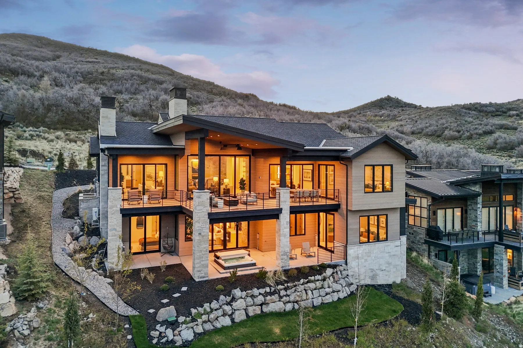 Back of house at dusk with a mountain view, stone siding, a patio area, a balcony, and a chimney