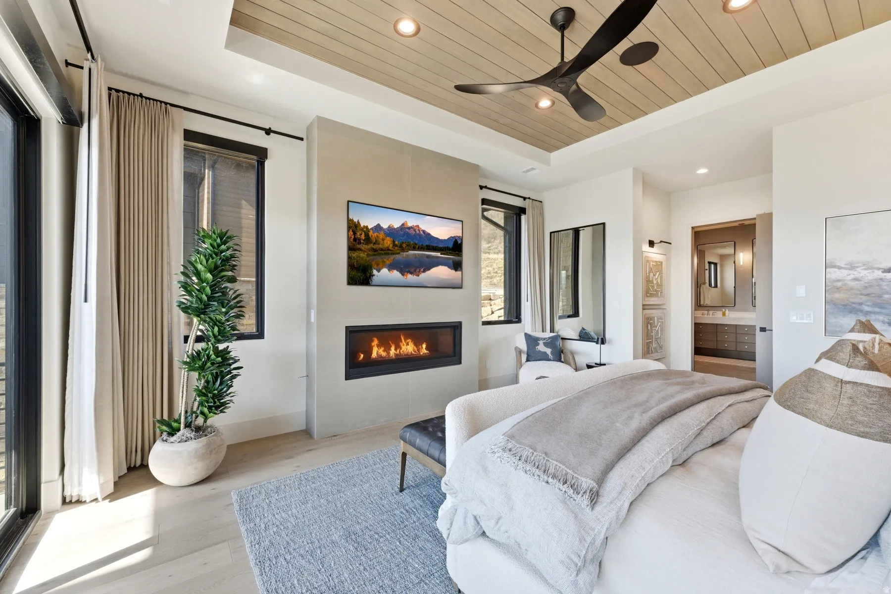 Bedroom with a wood tray ceiling, light wood-style floors, connected bathroom, a large fireplace, and ceiling fan